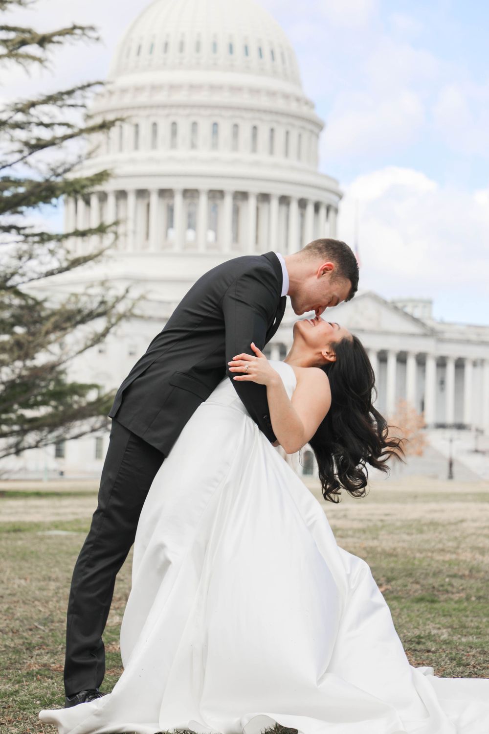 A romantic wedding photo of a couple in formal attire sharing a kiss in front of the US Capitol Building in Washington DC.