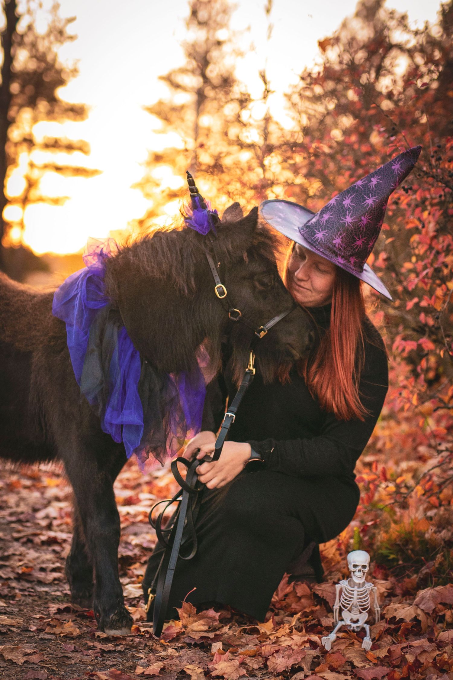 Witch in purple hat with decorated pony and small skeleton decoration in autumn forest at sunset.