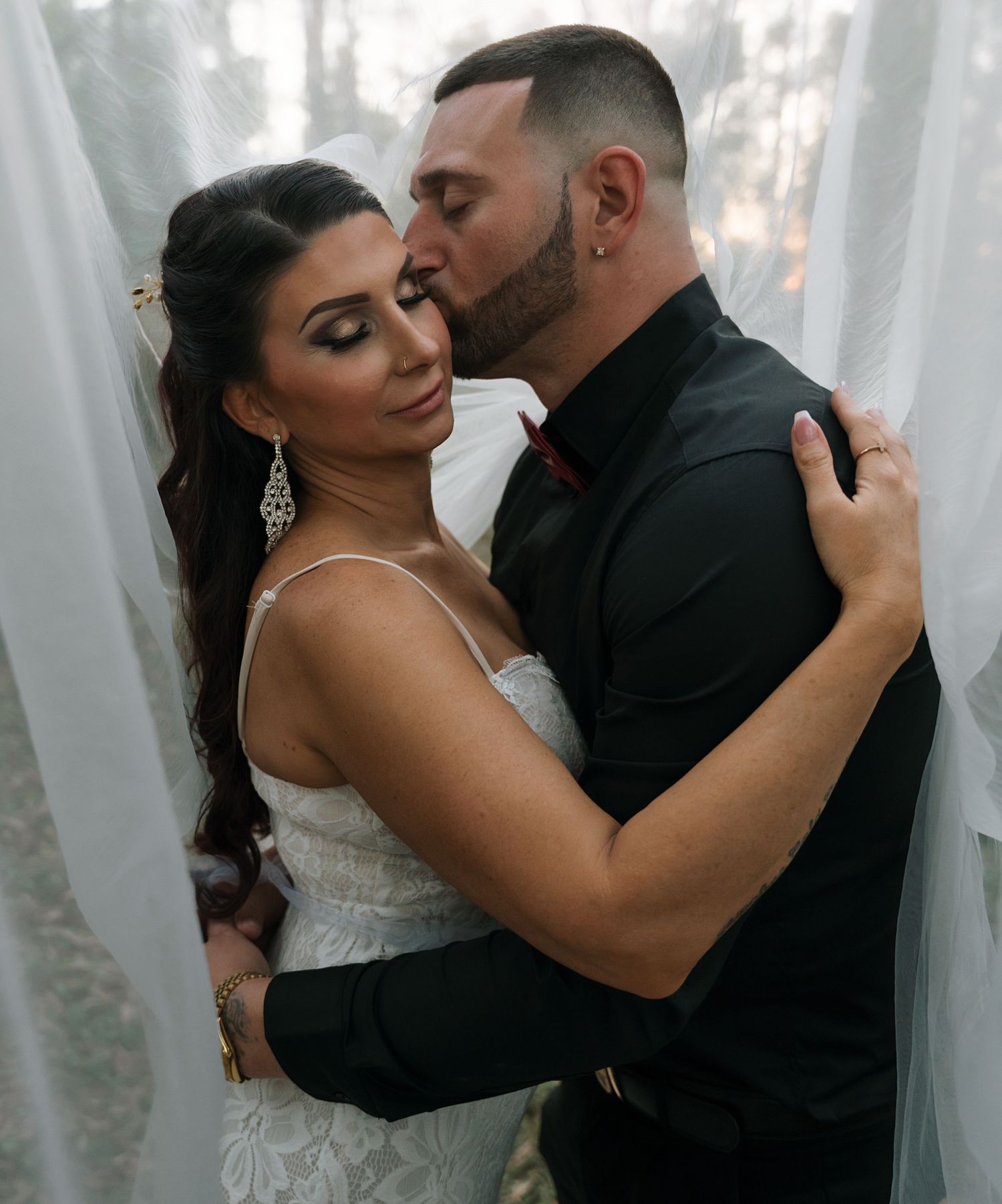A romantic couple shares an intimate moment during their wedding photoshoot among soft white curtains.
