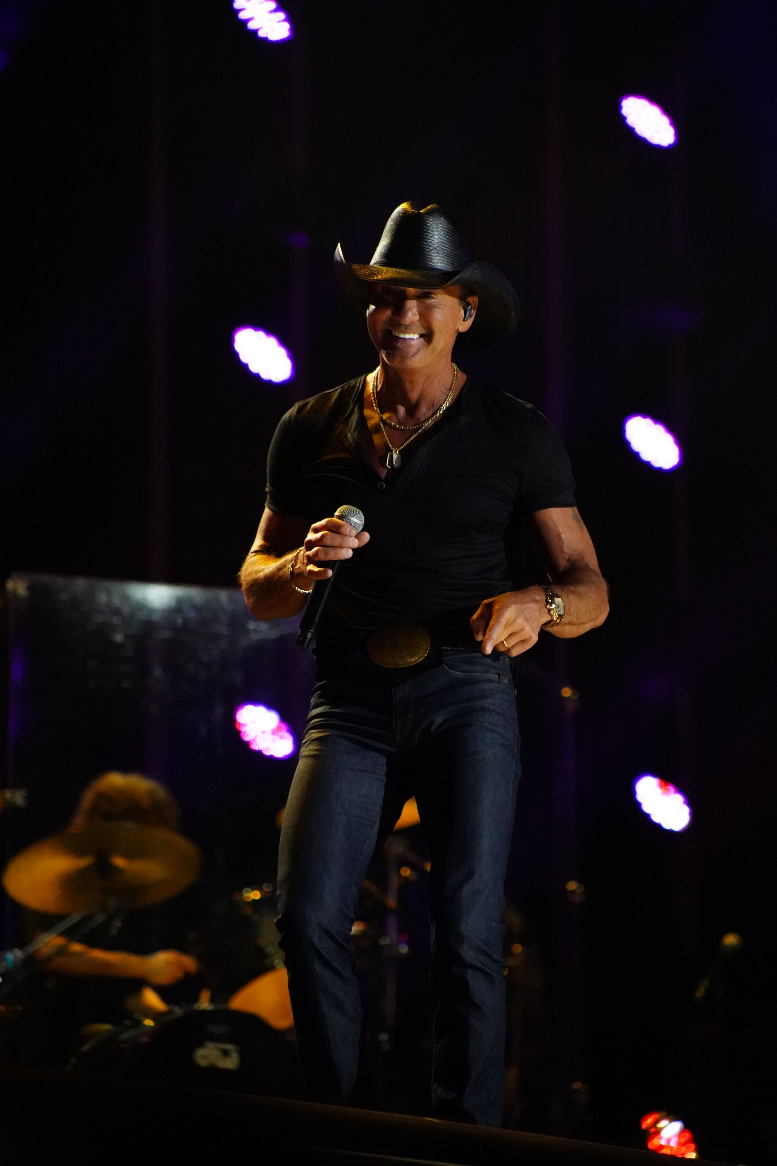 Concert performer in black cowboy hat singing on darkened stage with purple spotlights.