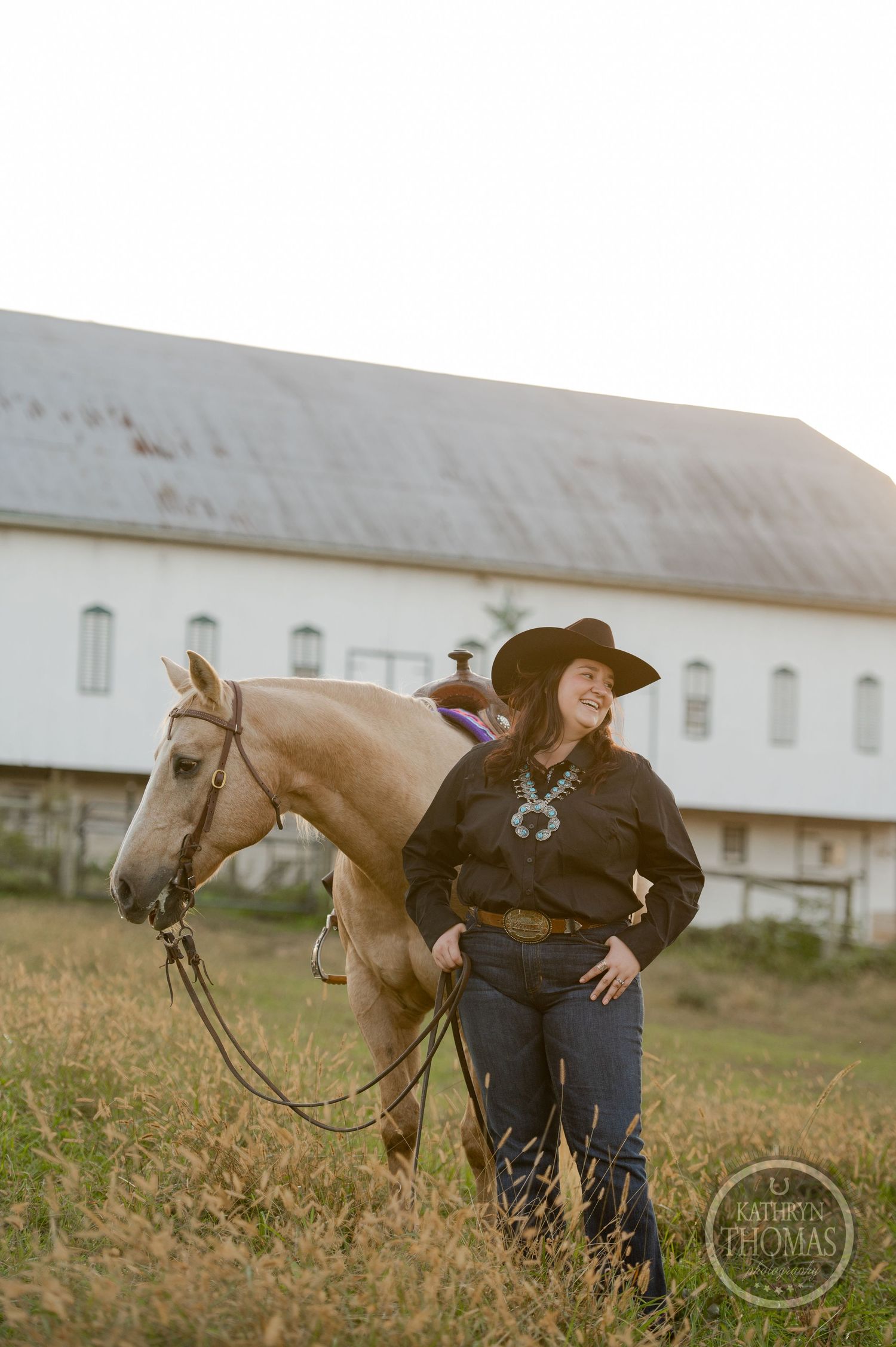 Woman in western attire stands beside a light-colored horse with a white barn in the background.