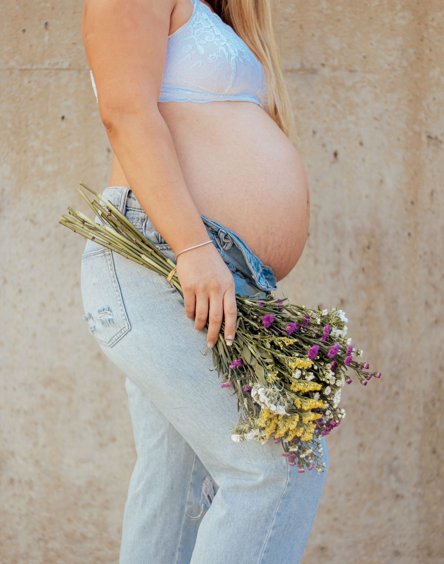 Side view of pregnant woman in light jeans and crop top holding dried wildflowers against beige wall.