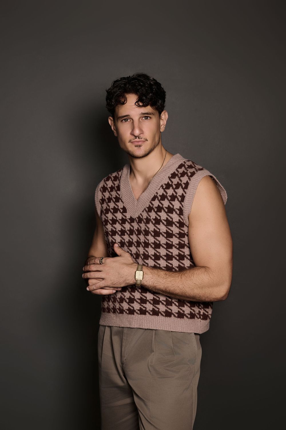 Man in houndstooth sweater vest and beige trousers posing confidently against a dark gray background.