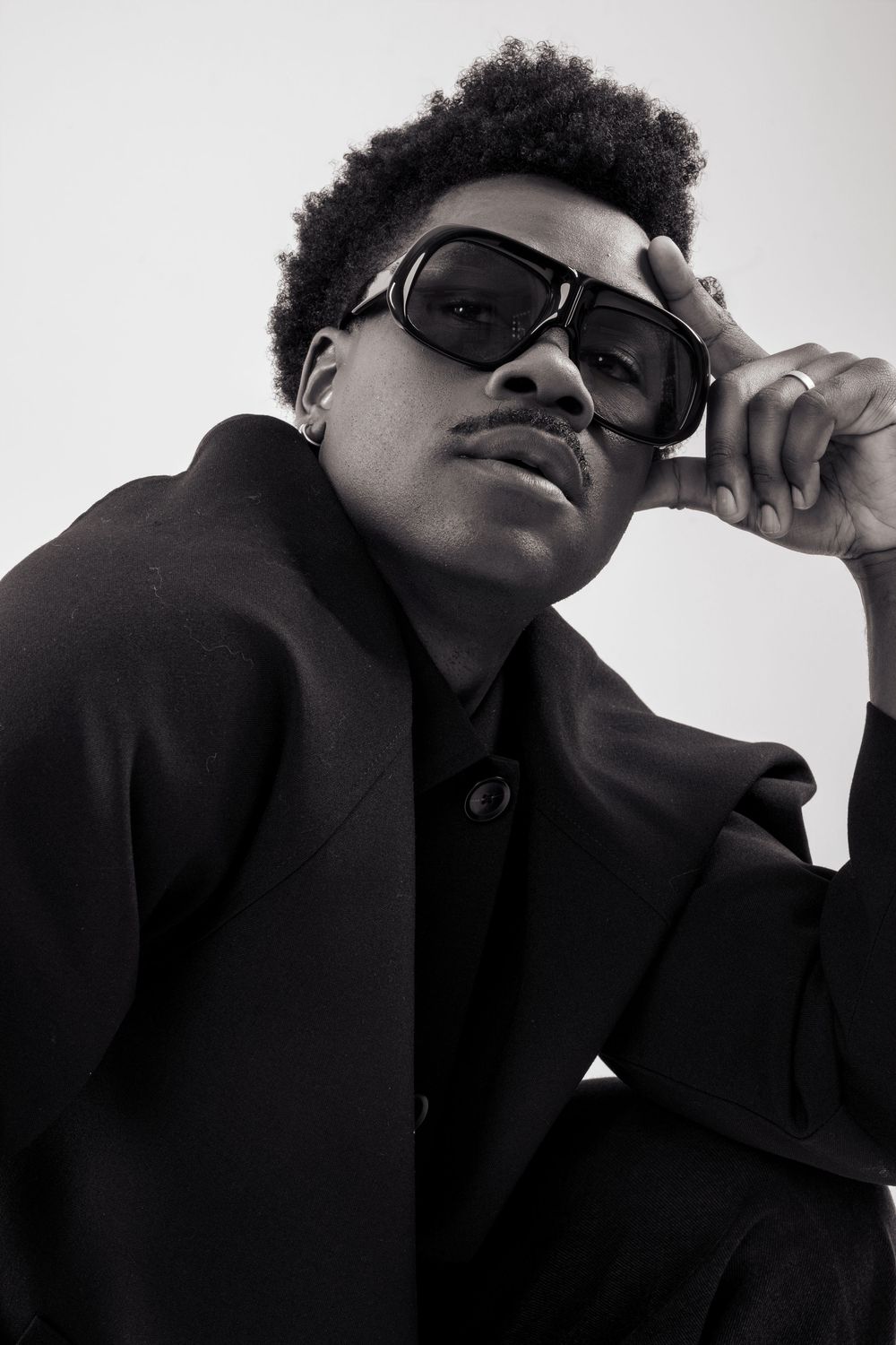 Black and white portrait of a stylish young man in a black blazer adjusting oversized sunglasses.