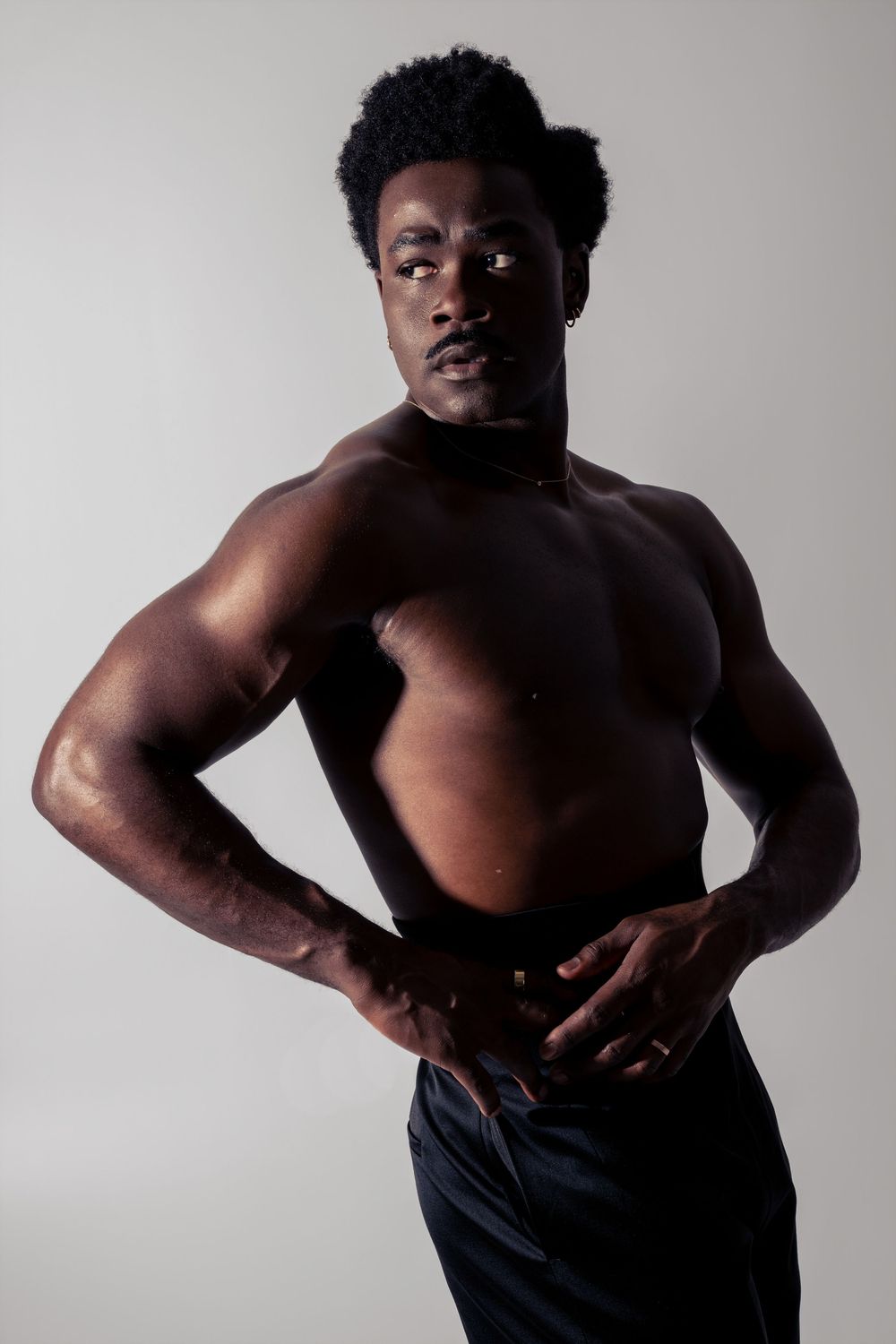Muscular shirtless Black man with afro hair posing confidently against a light gray studio background.