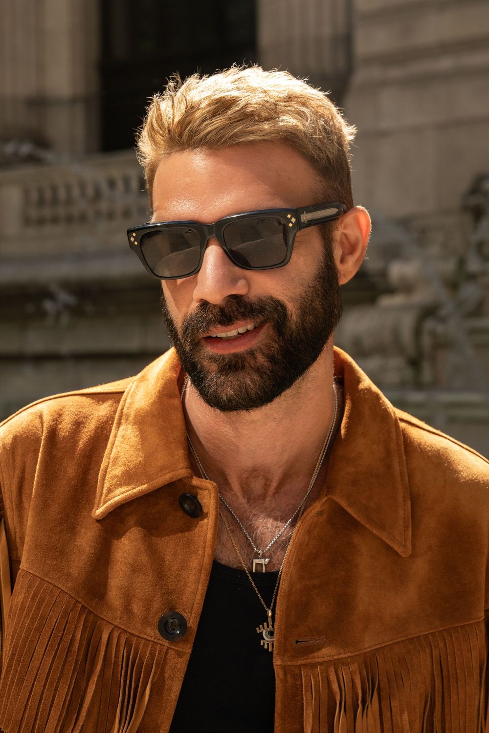Man with blonde hair and beard wearing black sunglasses and a brown suede fringe jacket outdoors.