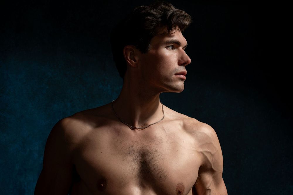 Muscular shirtless young man with gold necklace posing against dark blue background in dramatic studio lighting.
