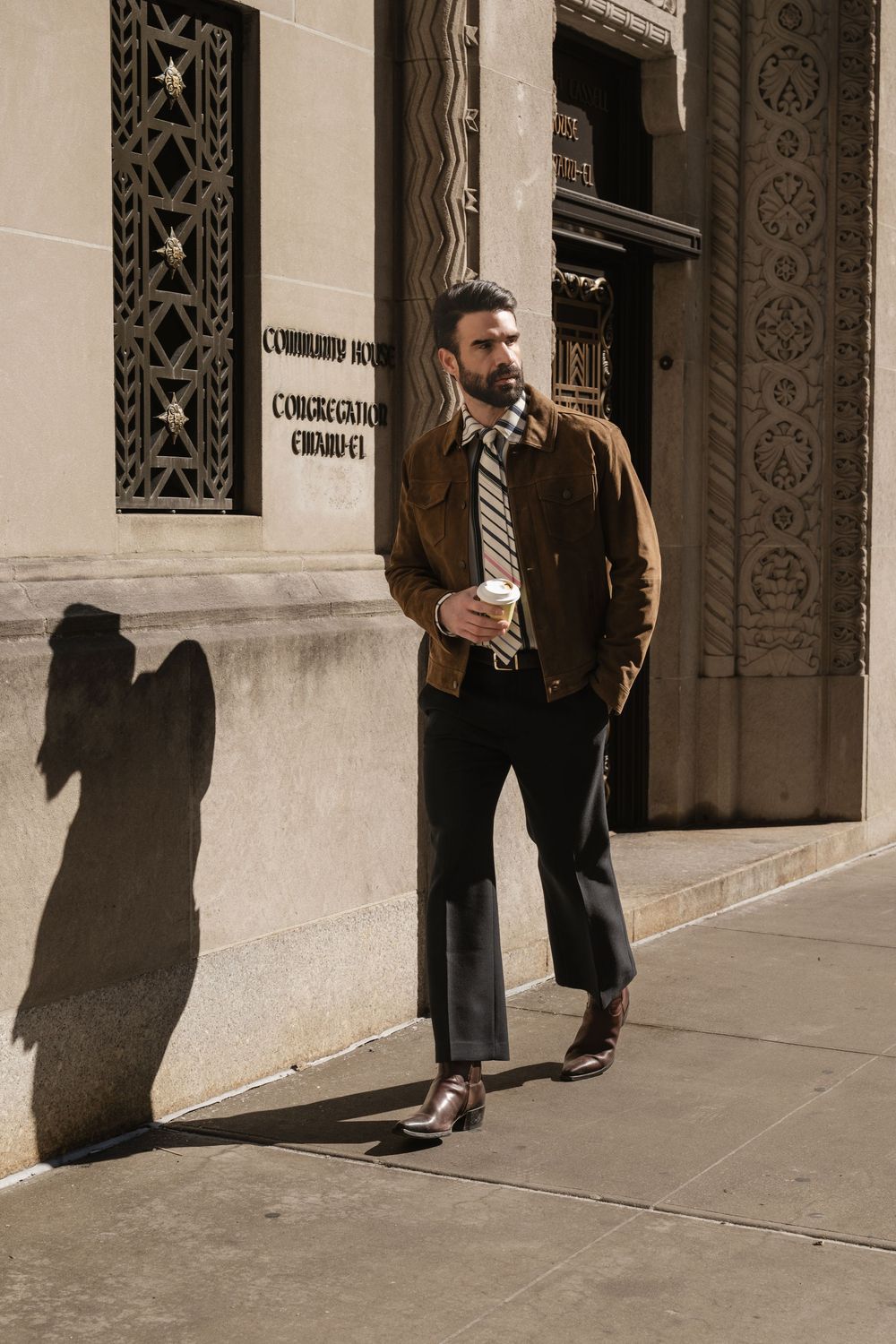 Fashion street photography of man in brown jacket walking past ornate architectural details.