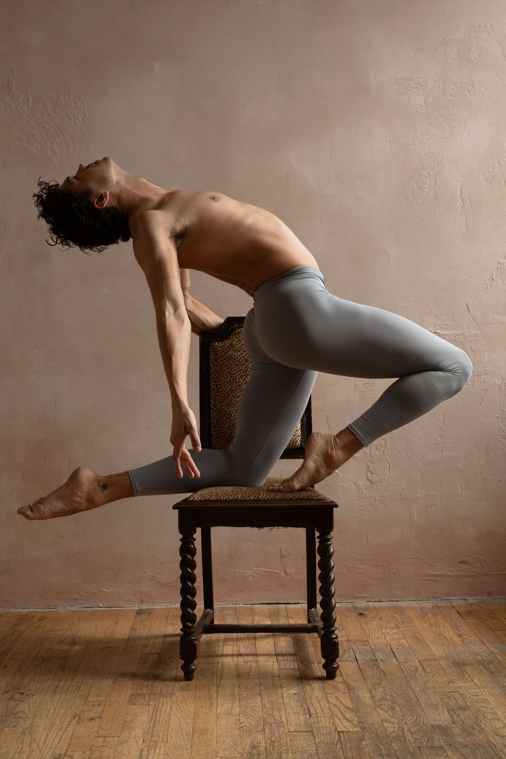 Male ballet dancer balancing on a wooden chair in gray leggings, arching back gracefully in a studio setting.