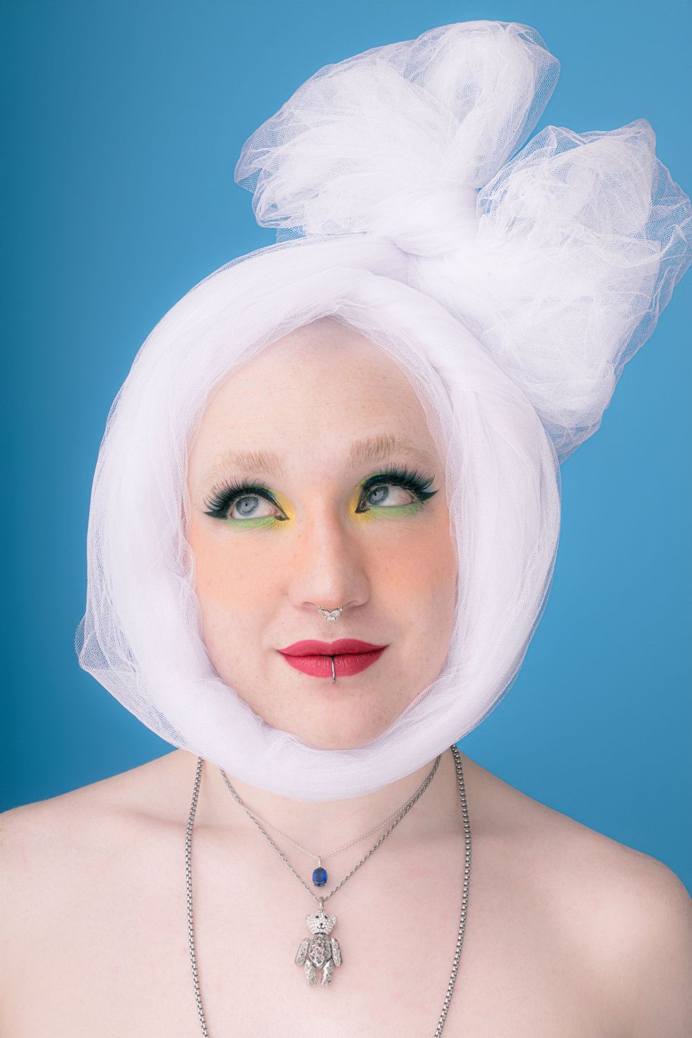 Artistic beauty portrait with white tulle head wrap, bold makeup, and hamsa necklace against blue background.