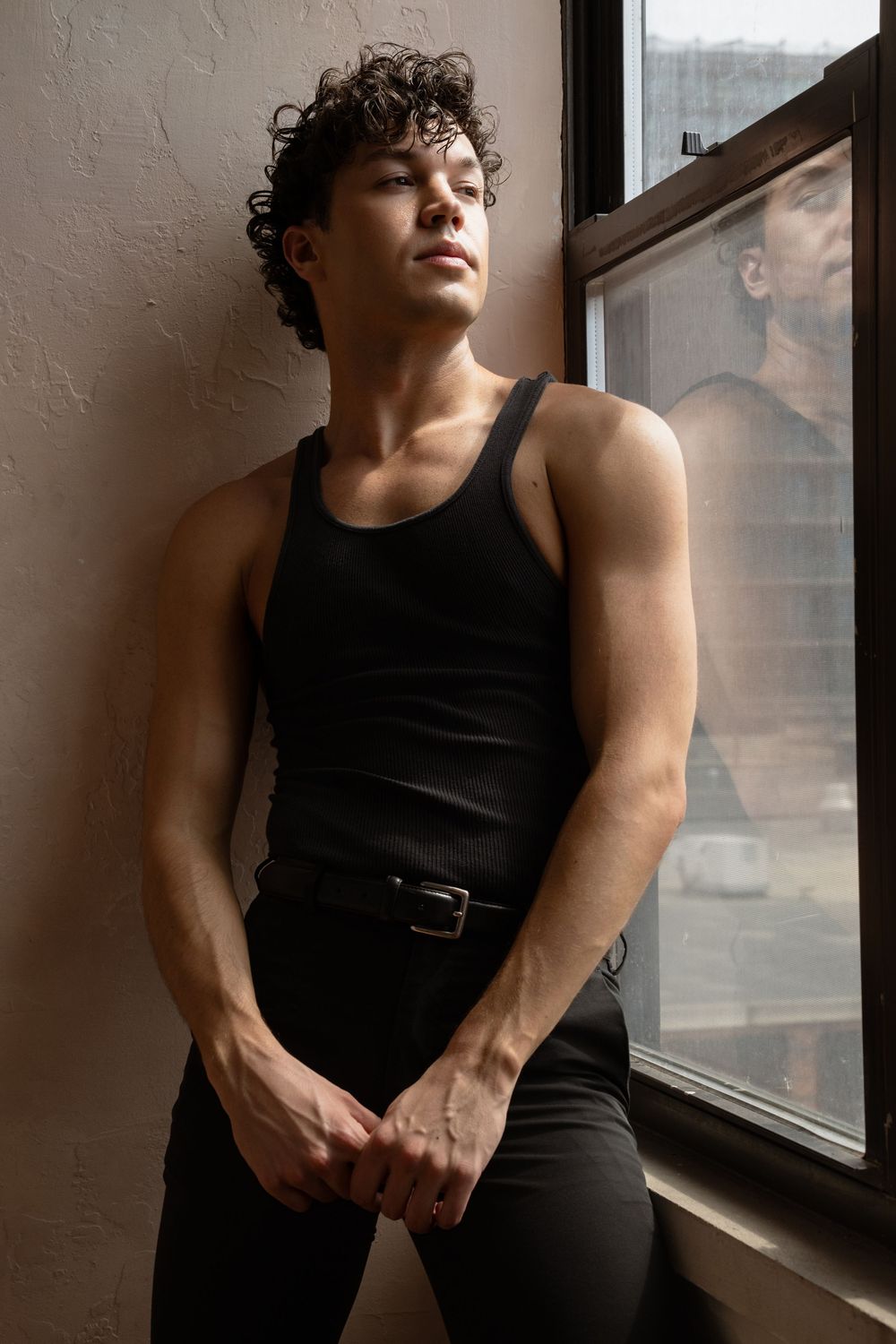 A muscular young man with curly hair wearing a black tank top and belt leans against a wall near a sunlit window.