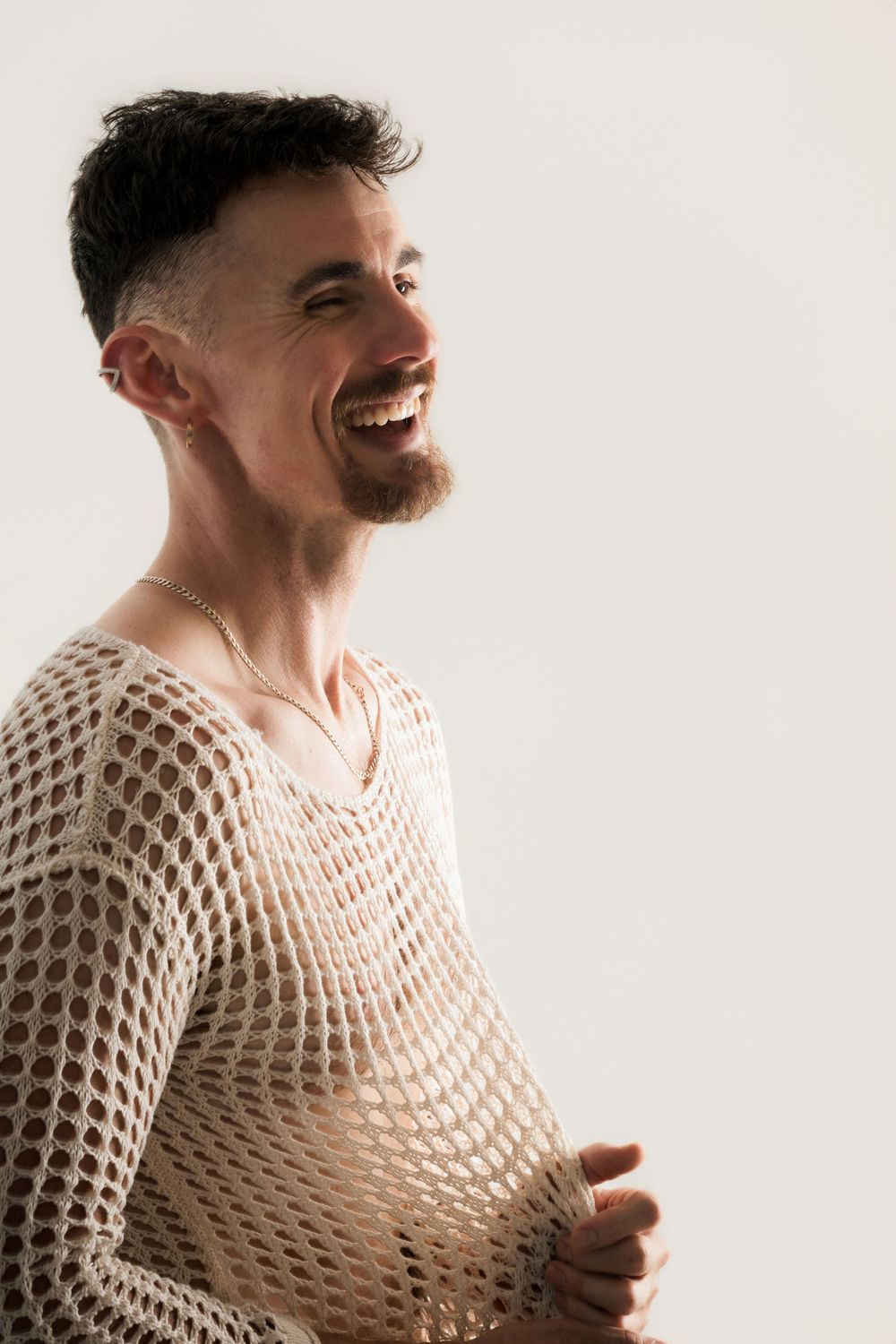 Smiling man with beard wearing a beige crochet mesh sweater and necklace, laughing against a white background.