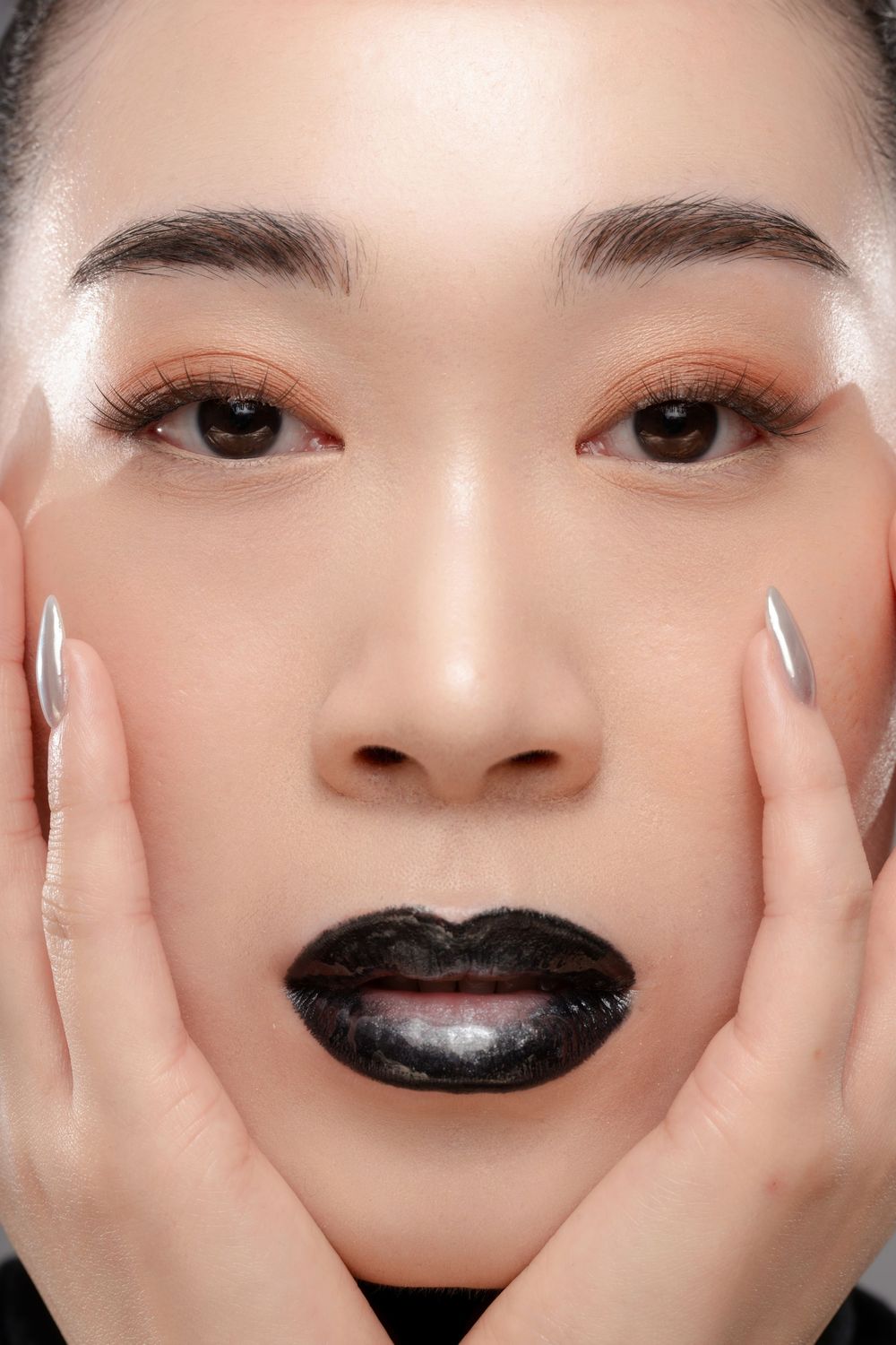 Close-up of a woman with dark black lips, metallic nails, warm eye makeup, and hands framing her face.