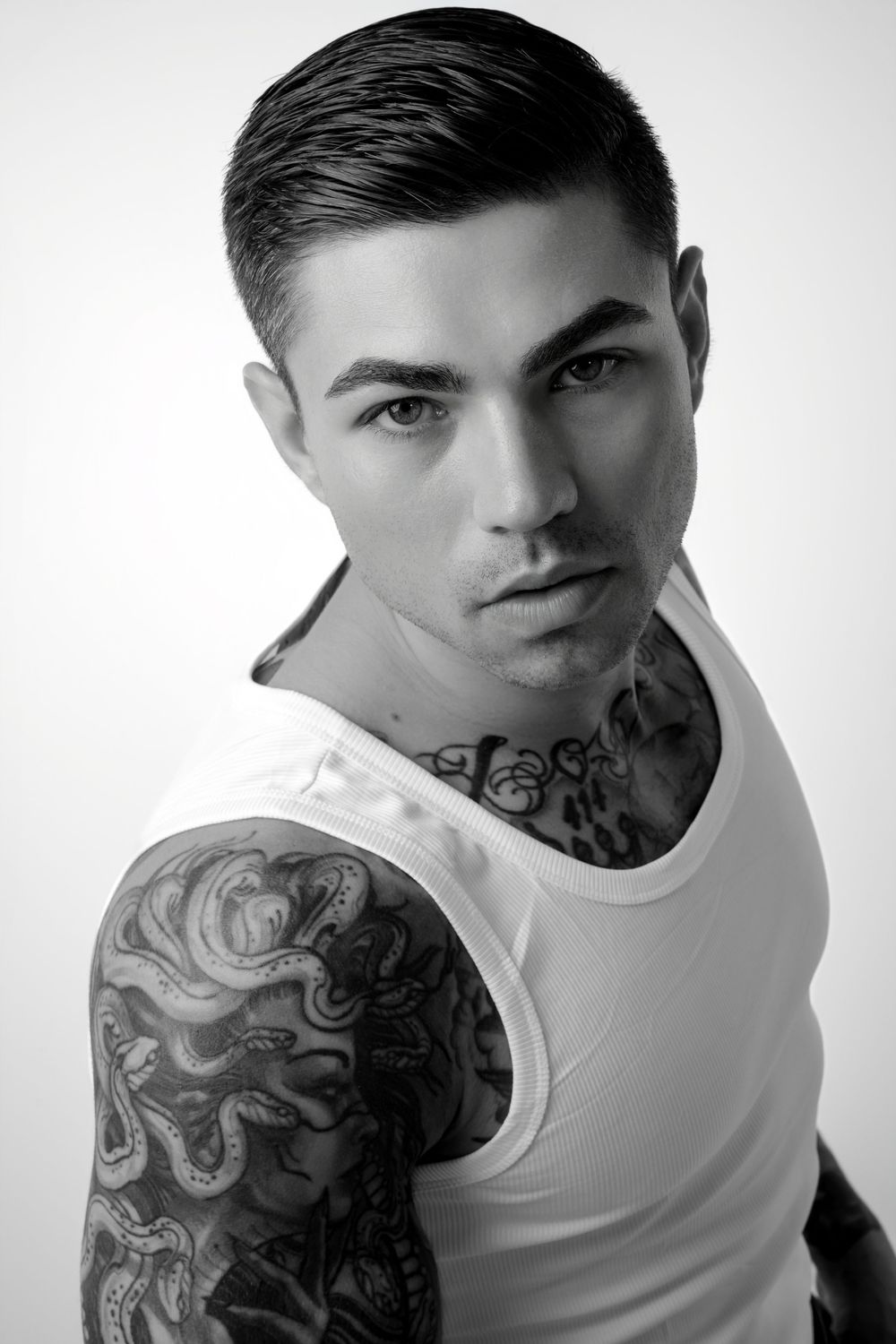Black and white portrait of a young tattooed man in a white tank top with sleeve and neck tattoos.