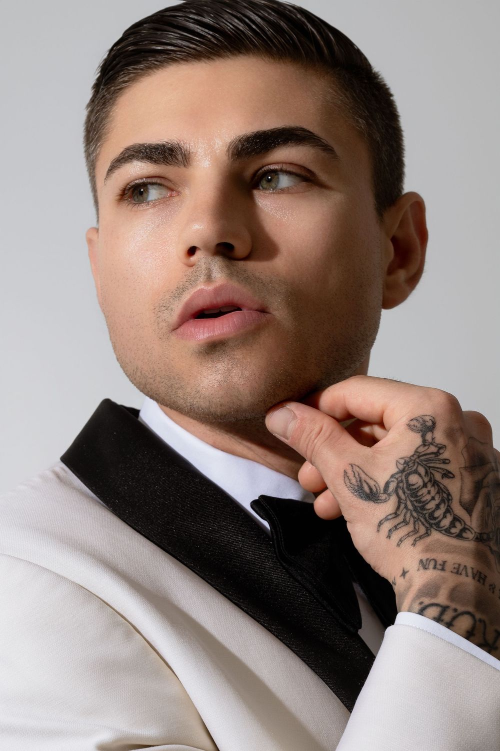 A stylish man in a white tuxedo with black lapels adjusts his bow tie, showcasing a scorpion tattoo on his hand.