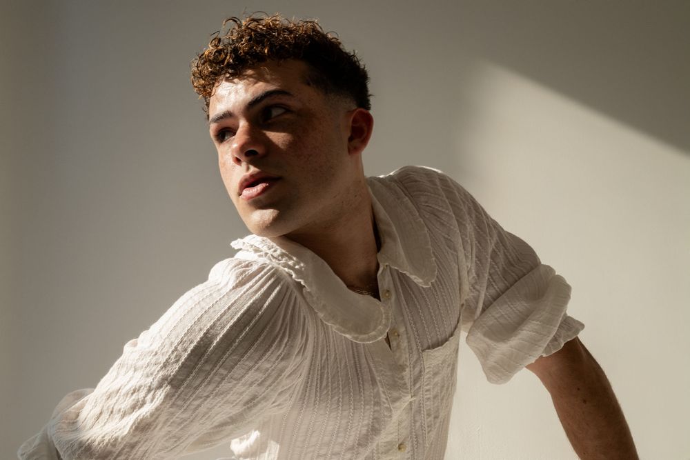 Young man in white textured short-sleeve shirt posing in soft natural light against a neutral wall.