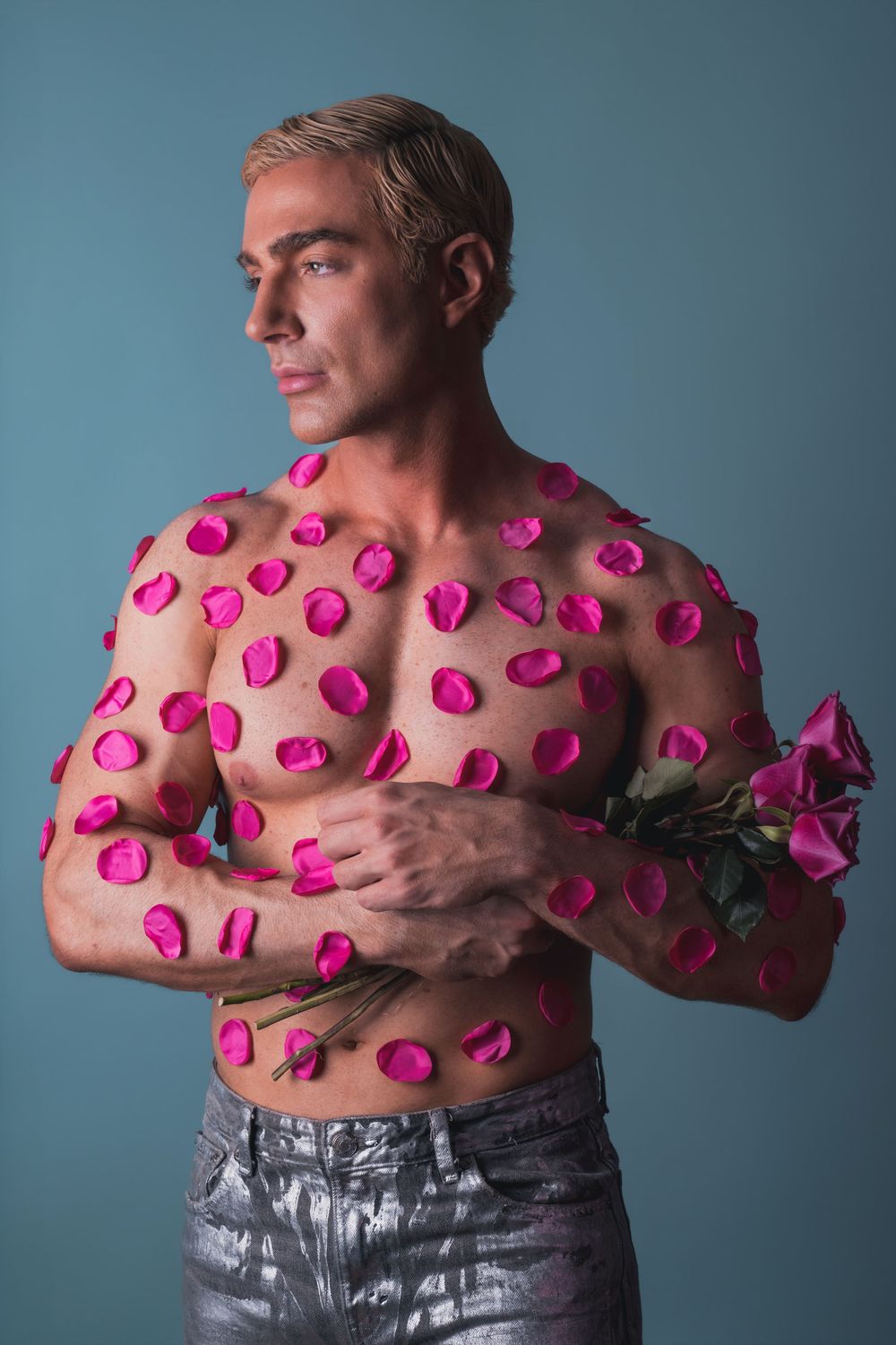 Shirtless muscular man with pink rose petals on torso holding roses, wearing silver pants against blue background.