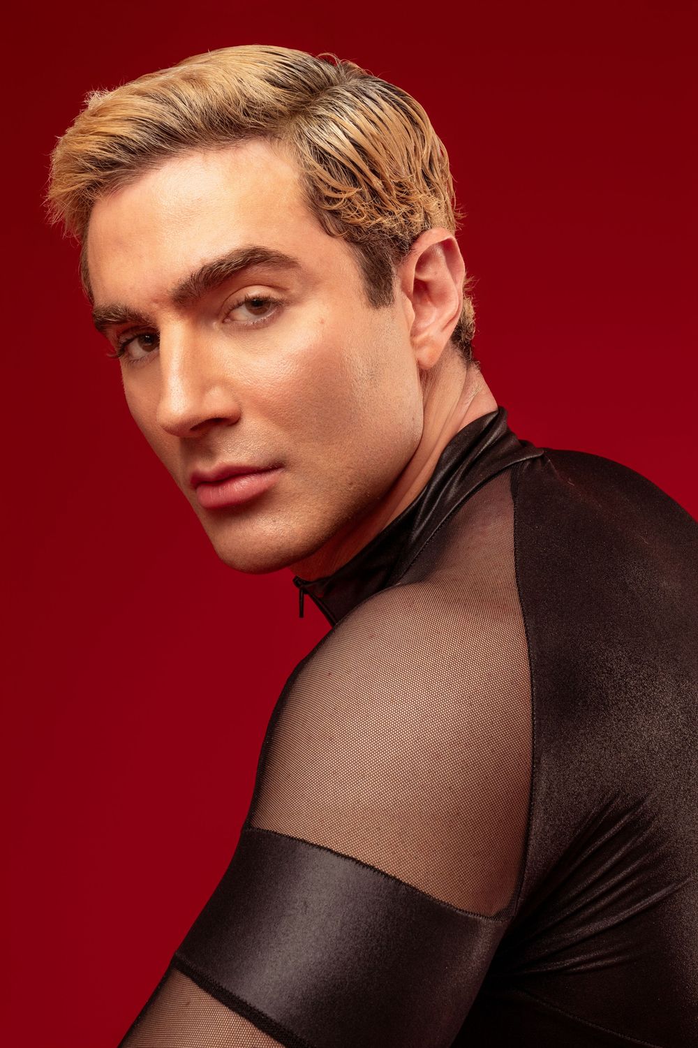 Fashion portrait against red background showing model in black mesh top with dramatic lighting and angles.