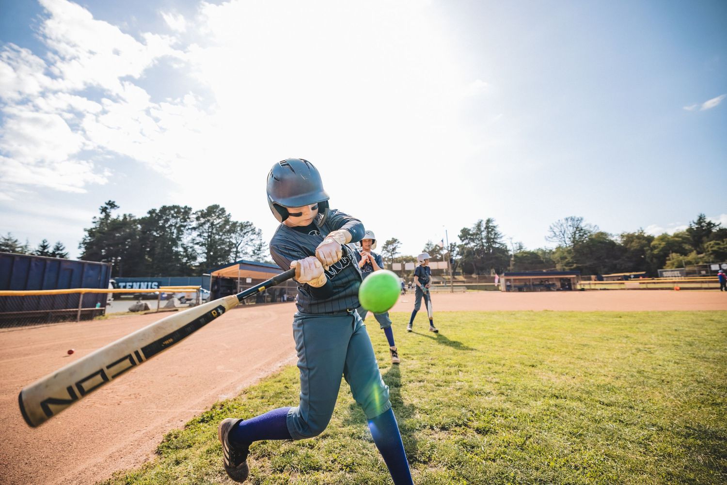 Baseball player in blue uniform takes a practice swing at batting cage on sunny day at baseball field.