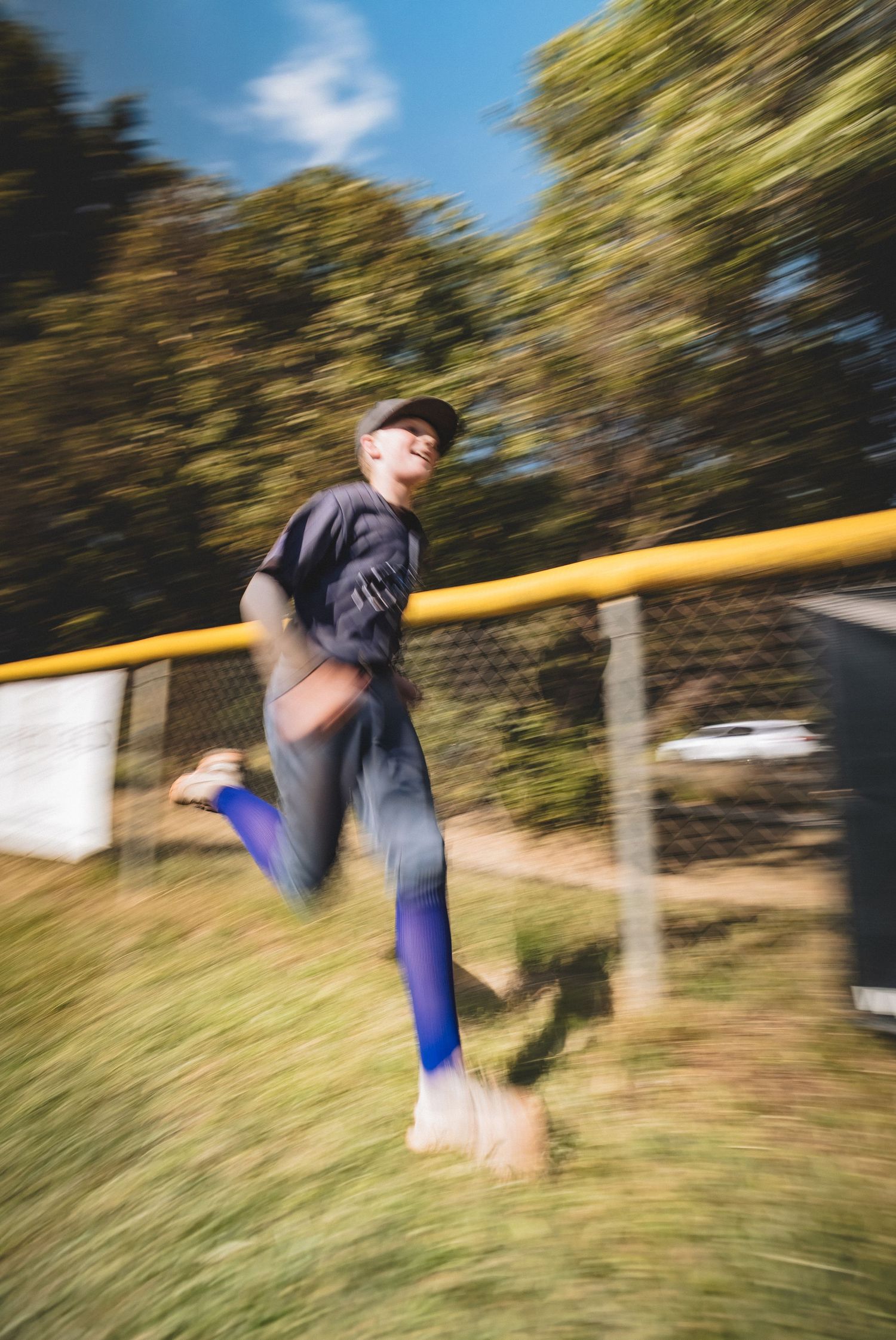 Motion blur photograph shows person in black shirt and blue socks jumping on grass with trees in background.