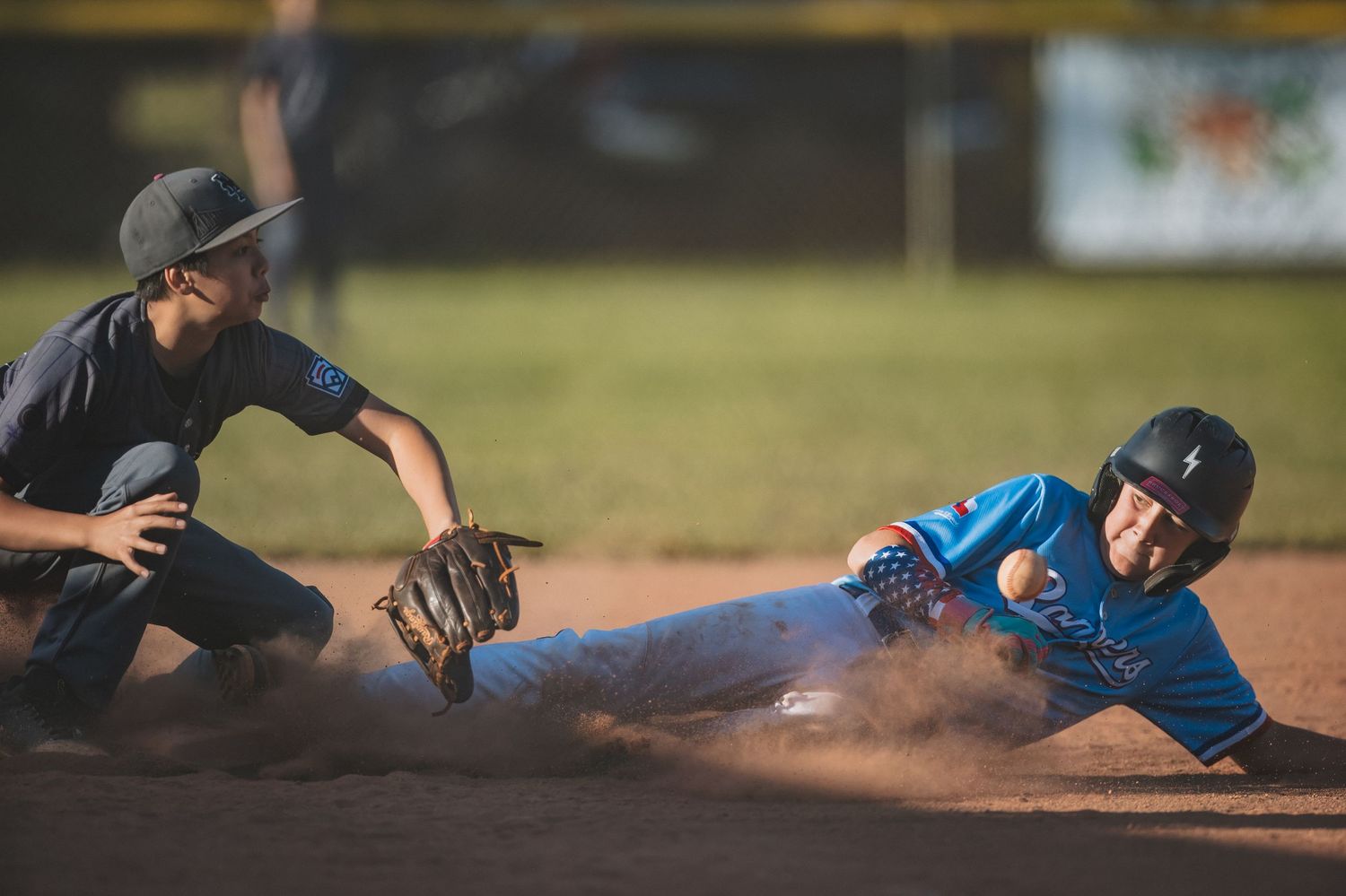 Baseball player in blue uniform slides into base as infielder attempts to make the tag while dust kicks up on the field.