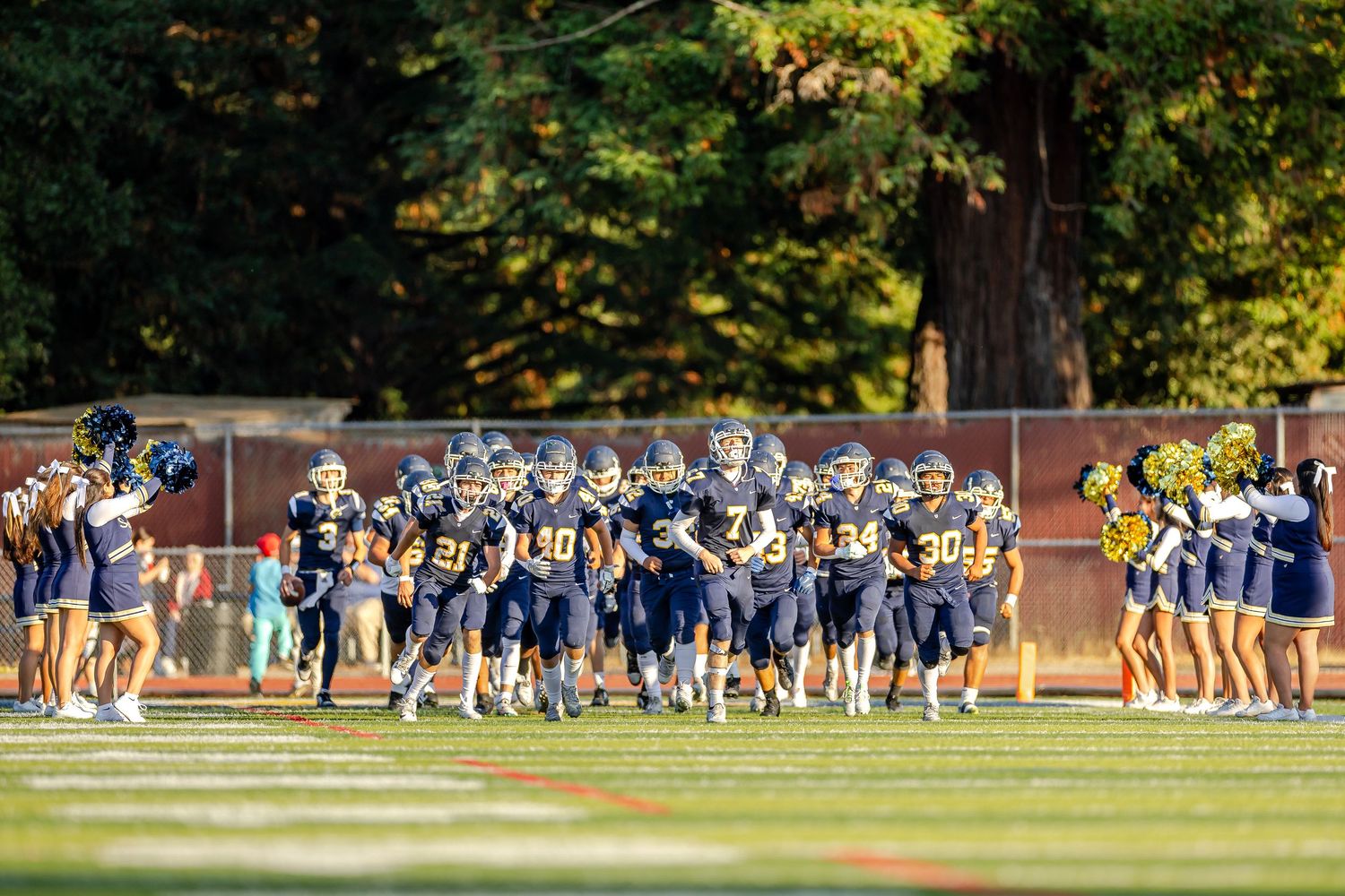 Football team players practicing drills and formations on a grass field bordered by autumn trees.