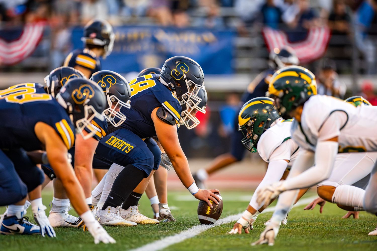 Football players line up at the line of scrimmage during a football game with offensive and defensive players ready to snap.