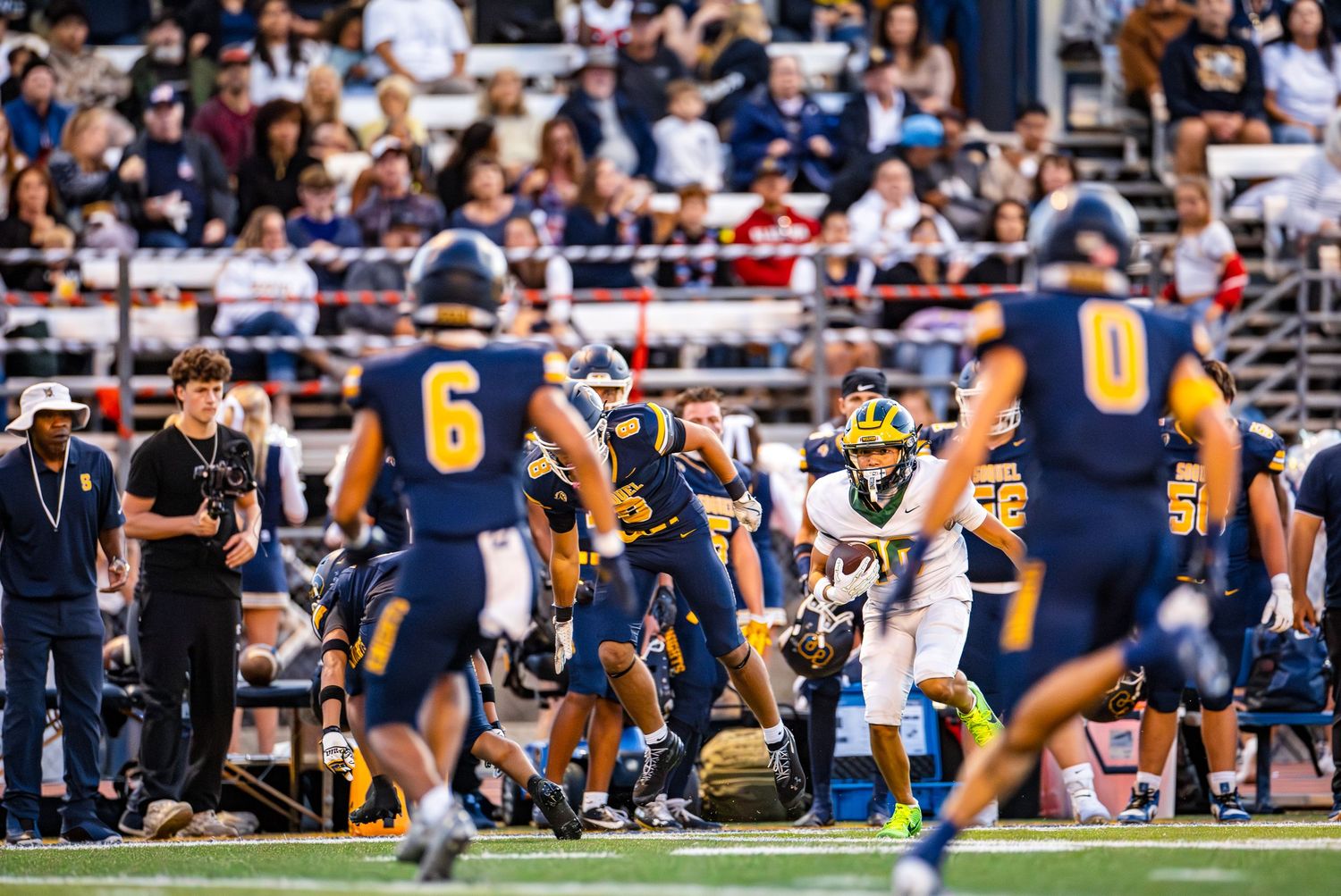 Football players crouch in a three-point stance at the line of scrimmage during a game between navy and yellow uniformed teams.