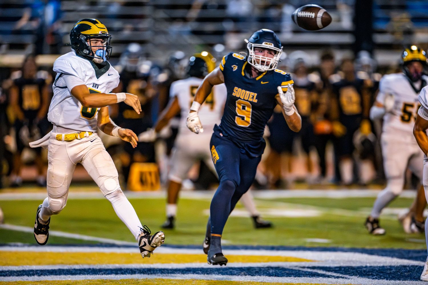 A high school football game shows players in motion with a football spiraling through the air during a night game.