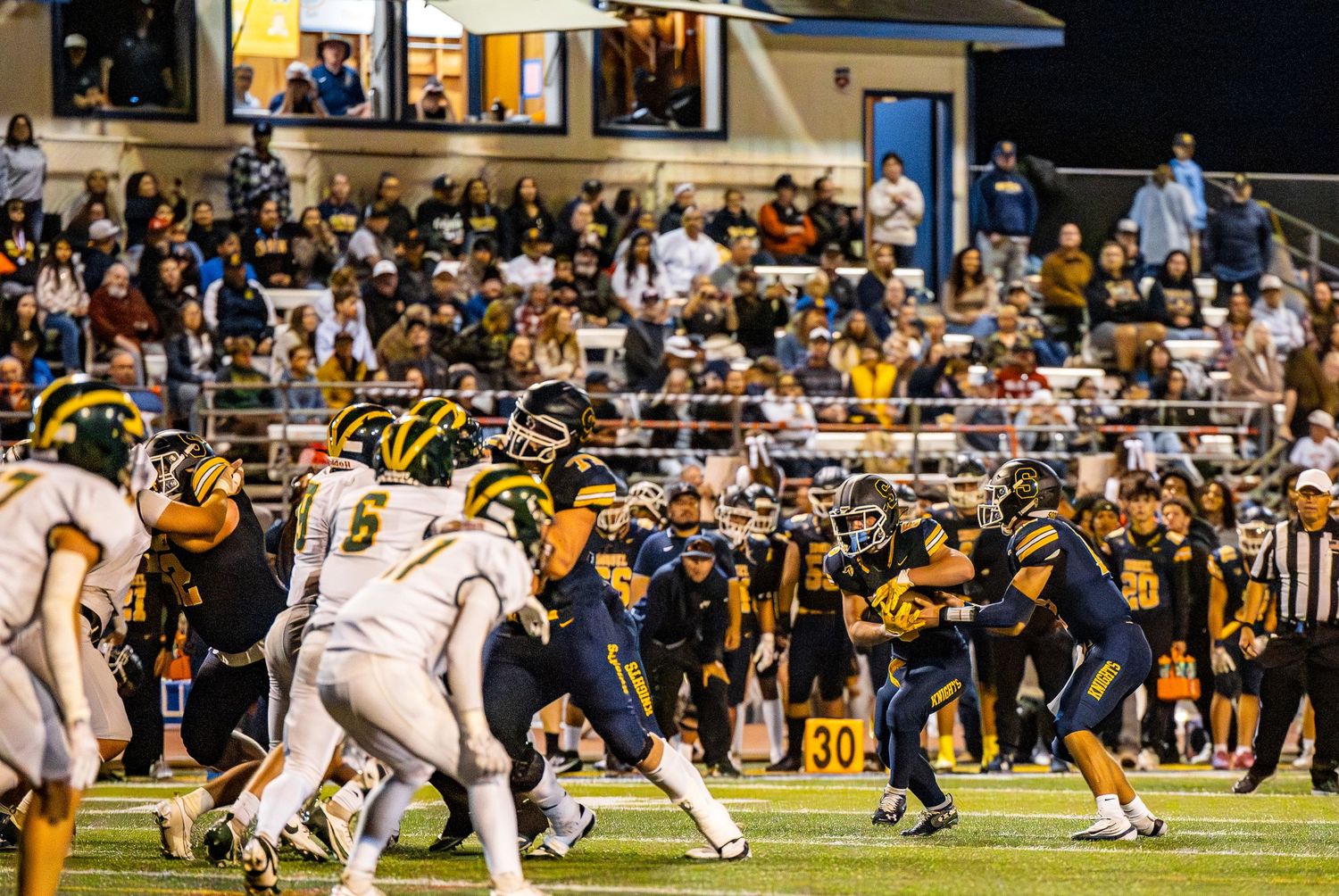 High school football teams face off during a field goal attempt at a night game with crowded bleachers in the background.