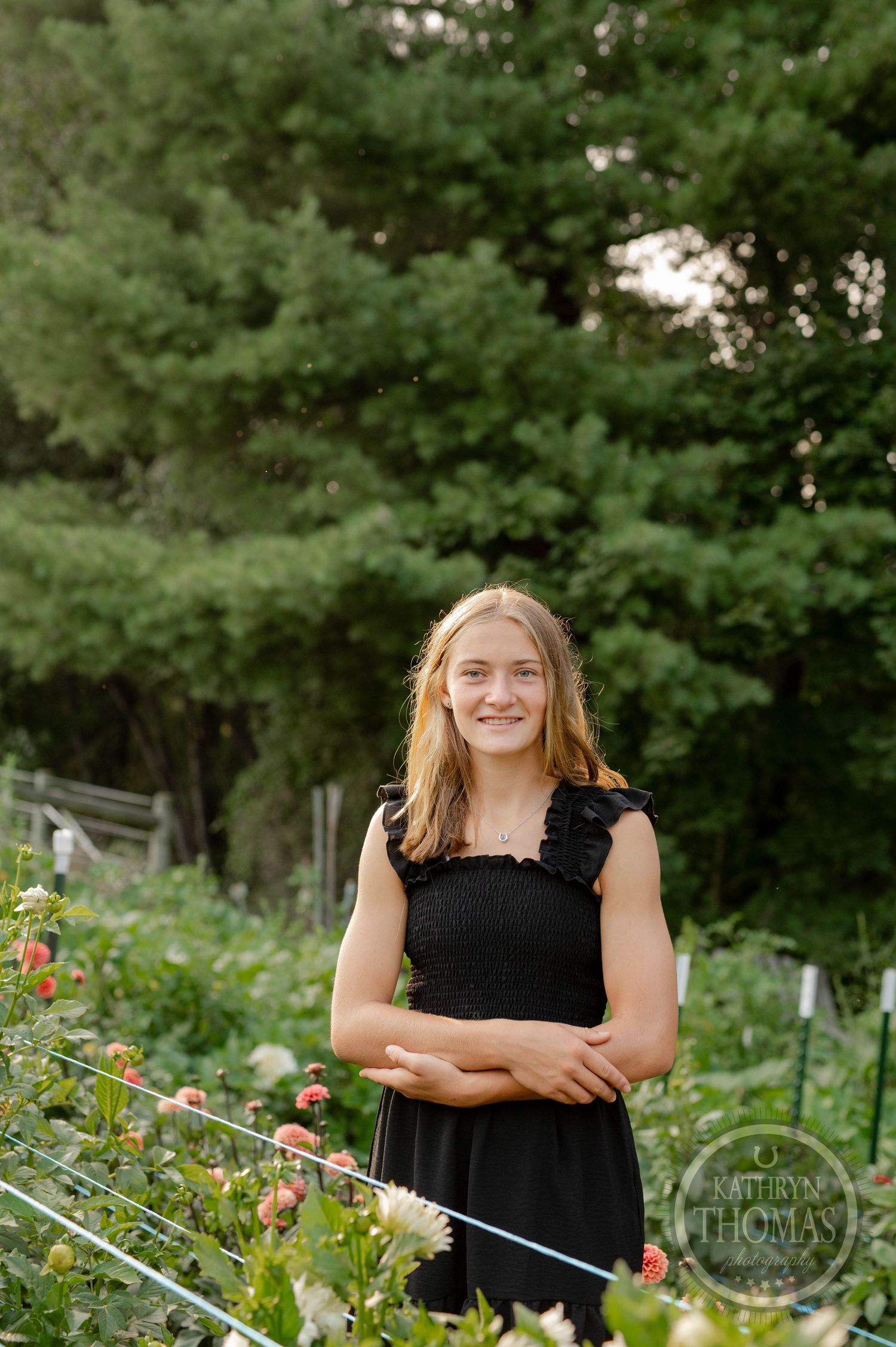 Woman in black sleeveless dress poses in front of garden with blooming flowers and green foliage.