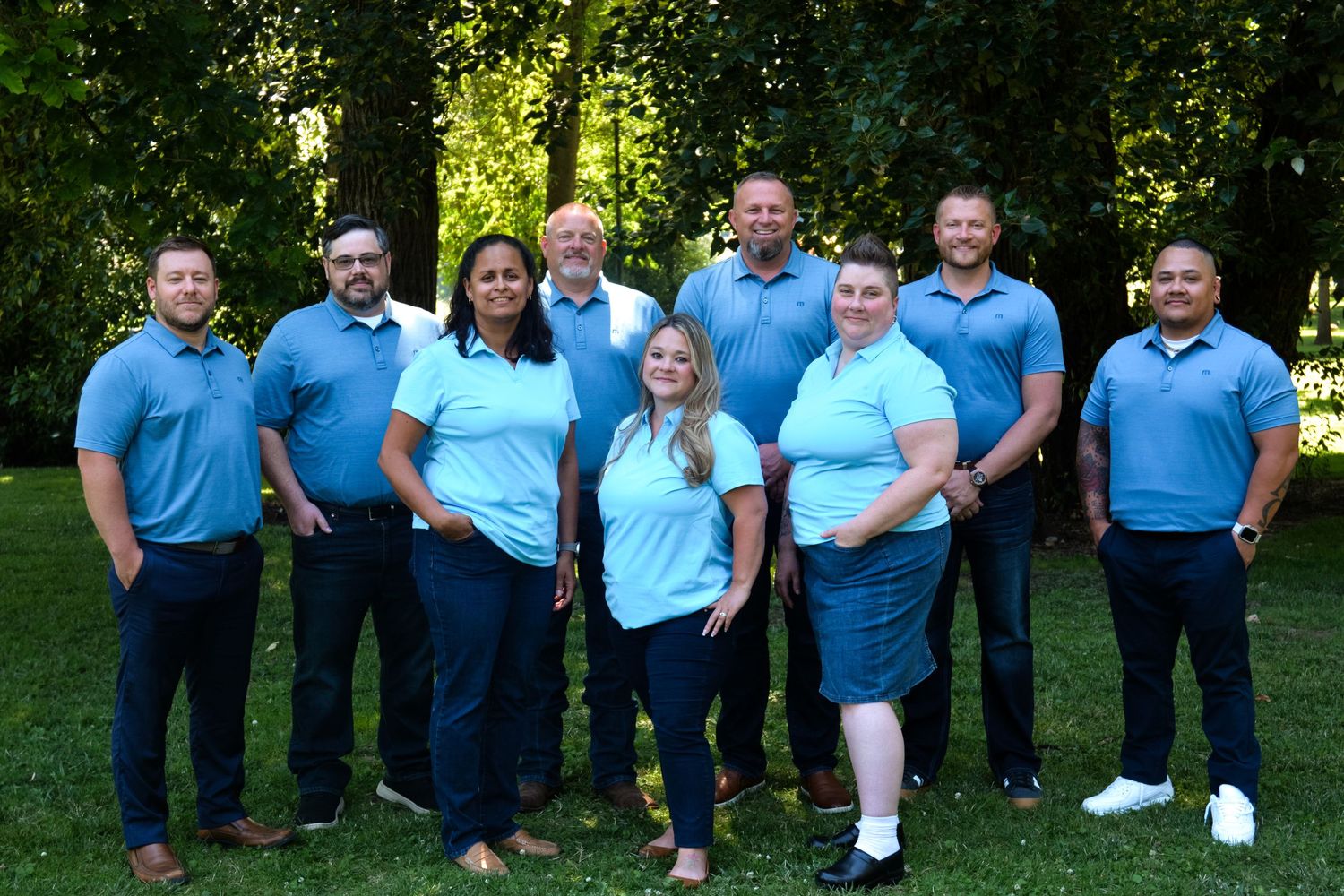 A group photo of team members wearing matching light blue polo shirts standing together outdoors in a park setting.