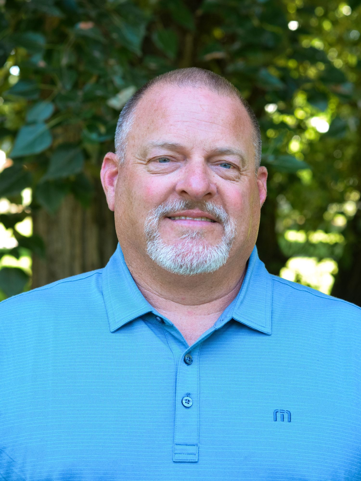 Outdoor portrait of someone in a blue polo shirt smiling against a green leafy background on a sunny day.