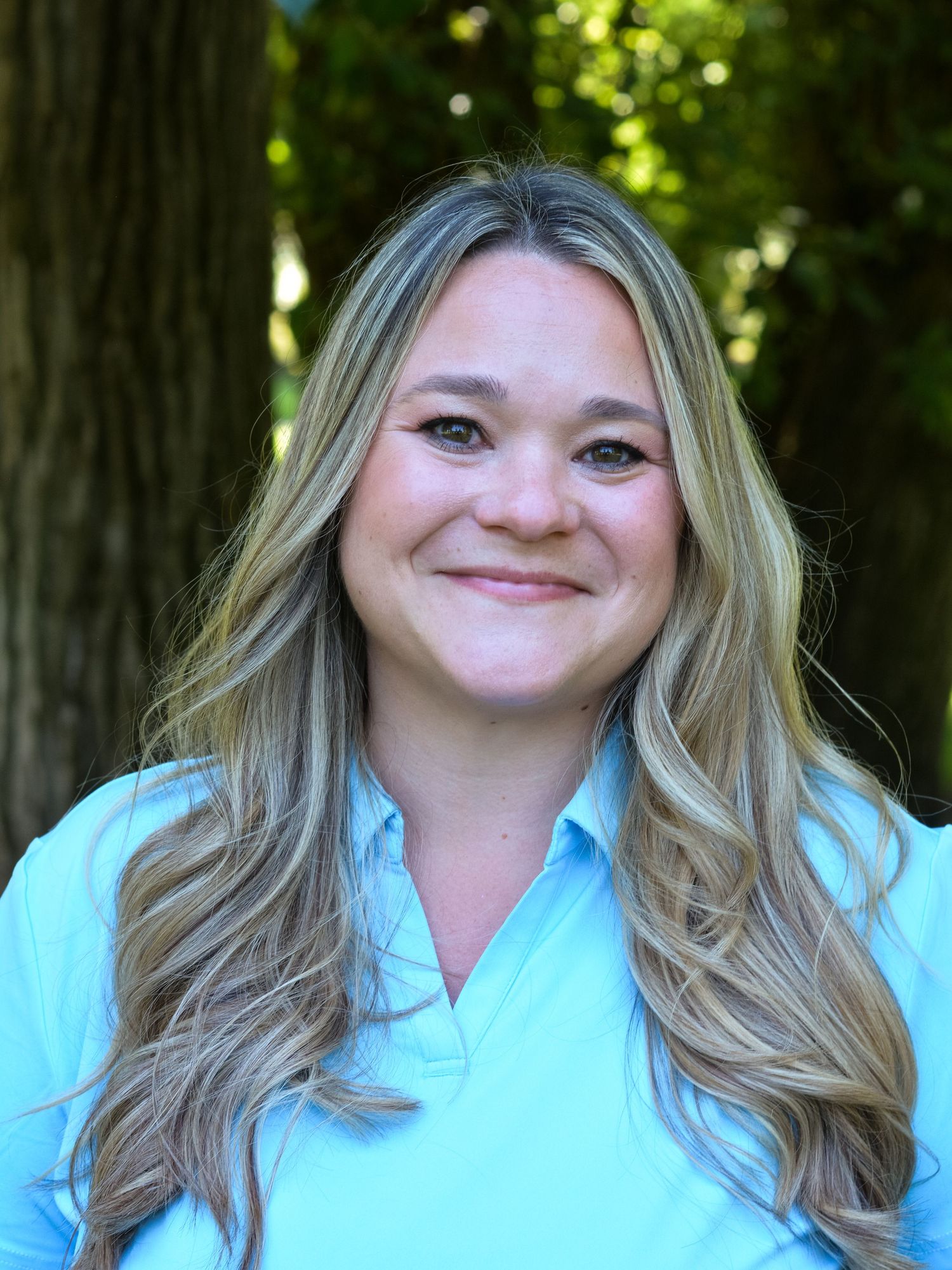 Professional headshot of person in light blue shirt against green outdoor background.