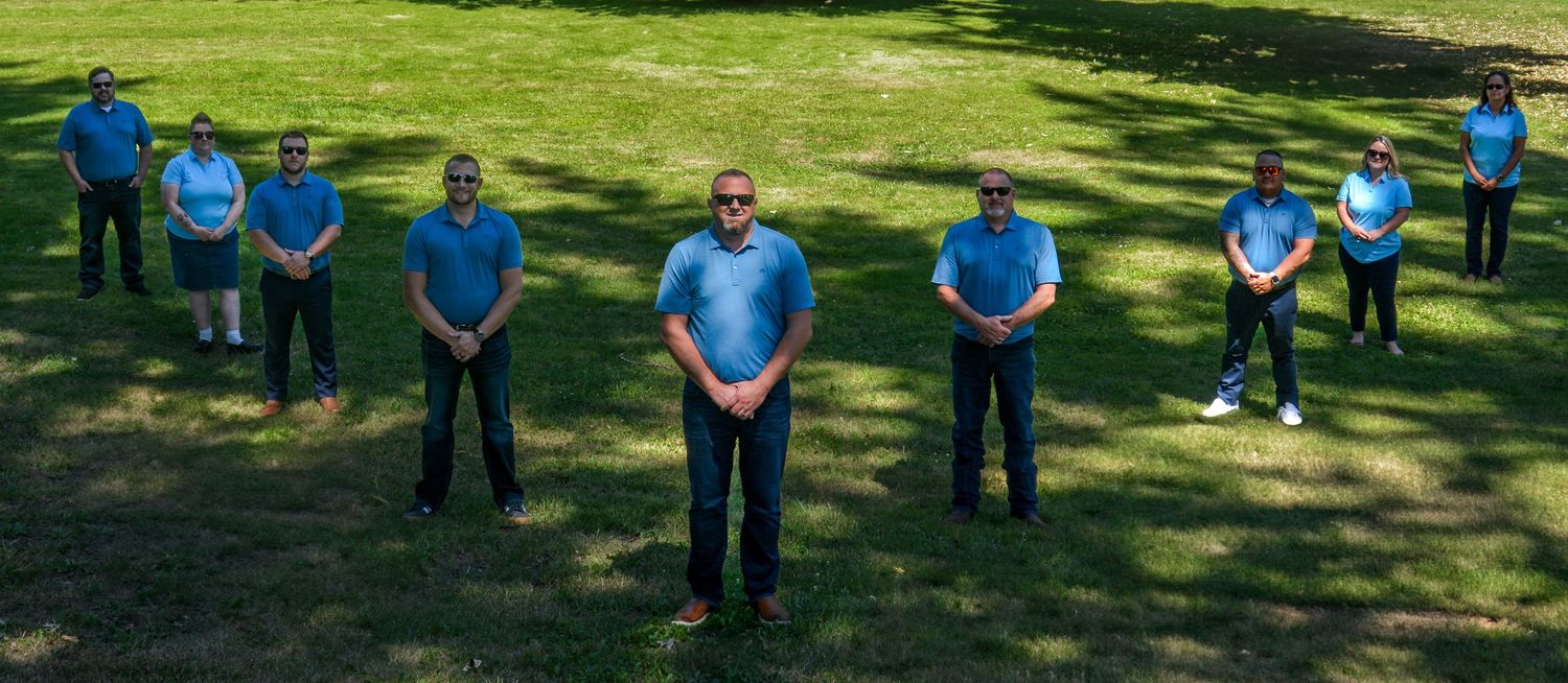 Team members in matching blue shirts standing socially distanced on green grass lawn.