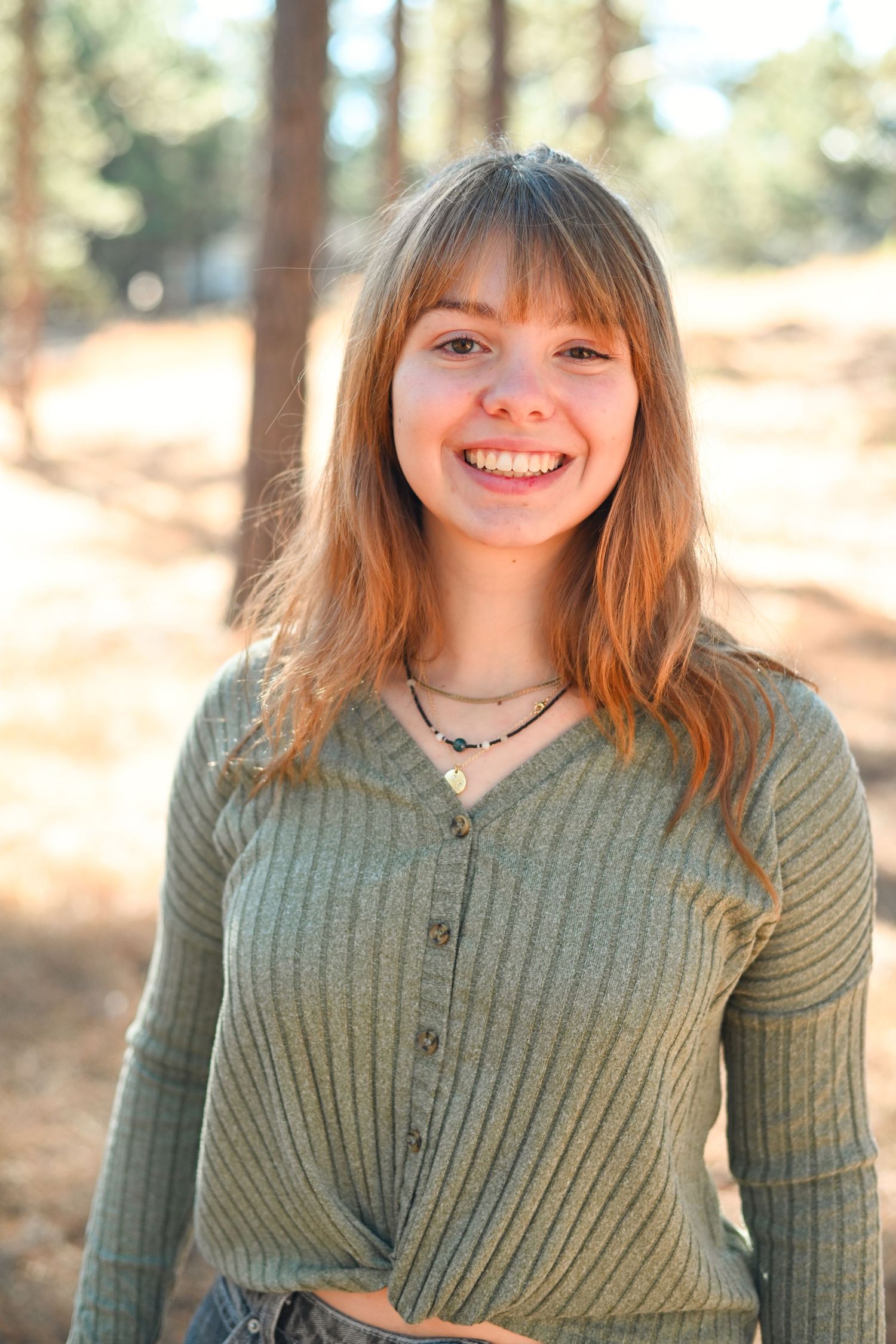 Smiling young woman with red hair wearing a green ribbed sweater outdoors among pine trees in warm sunlight.