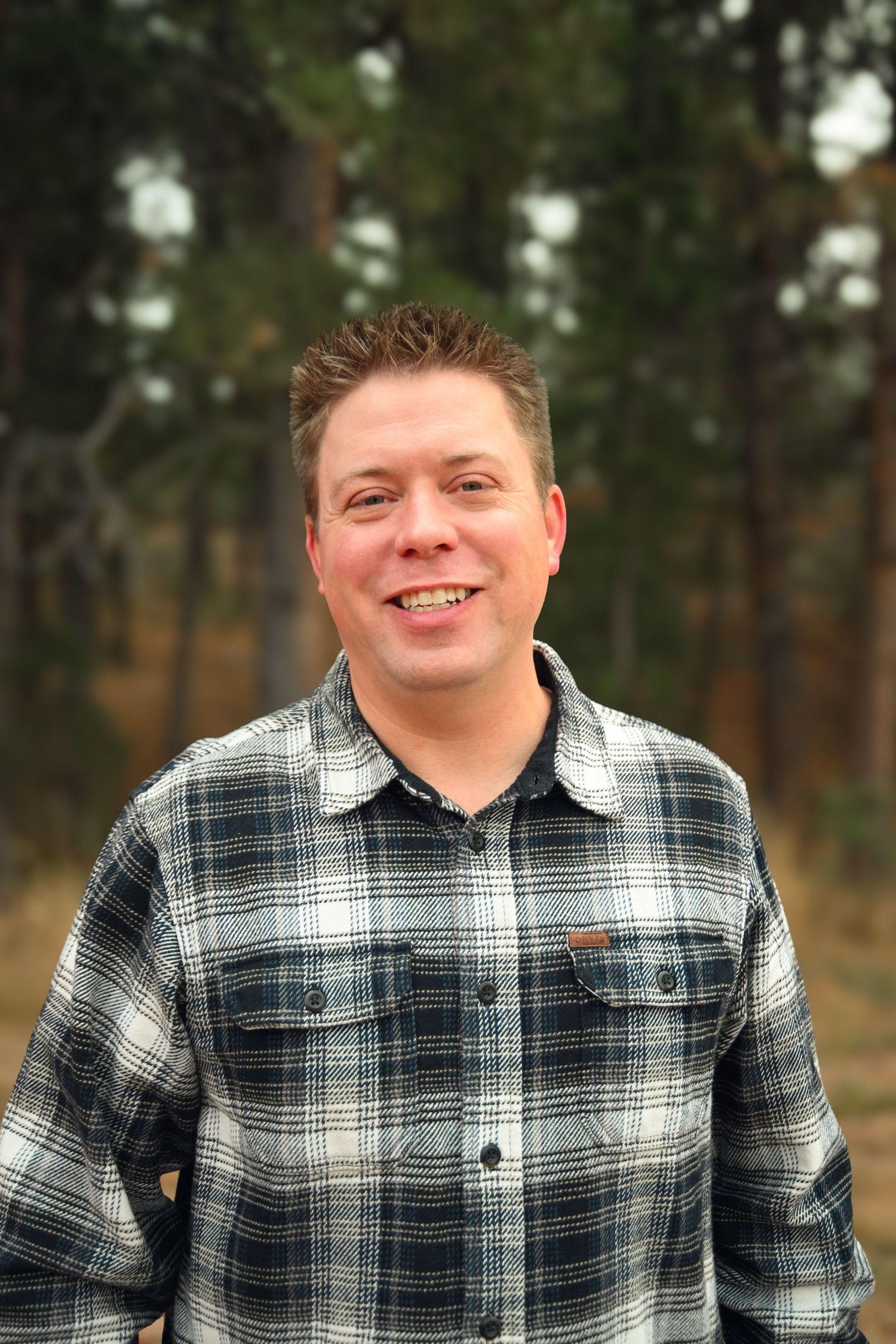 Smiling man in plaid flannel shirt standing outdoors in a forest setting with tall pine trees in background.