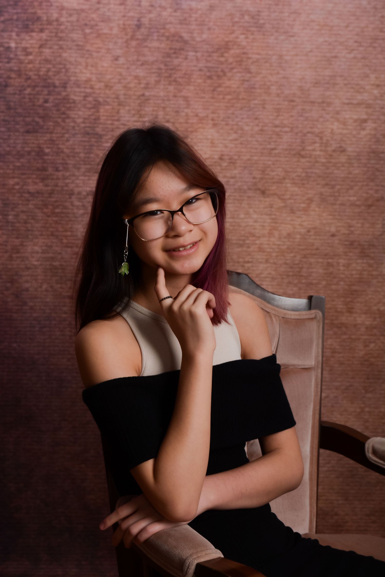 Portrait of a young Asian woman smiling in a black off-shoulder dress seated in a vintage chair against a brown backdrop.