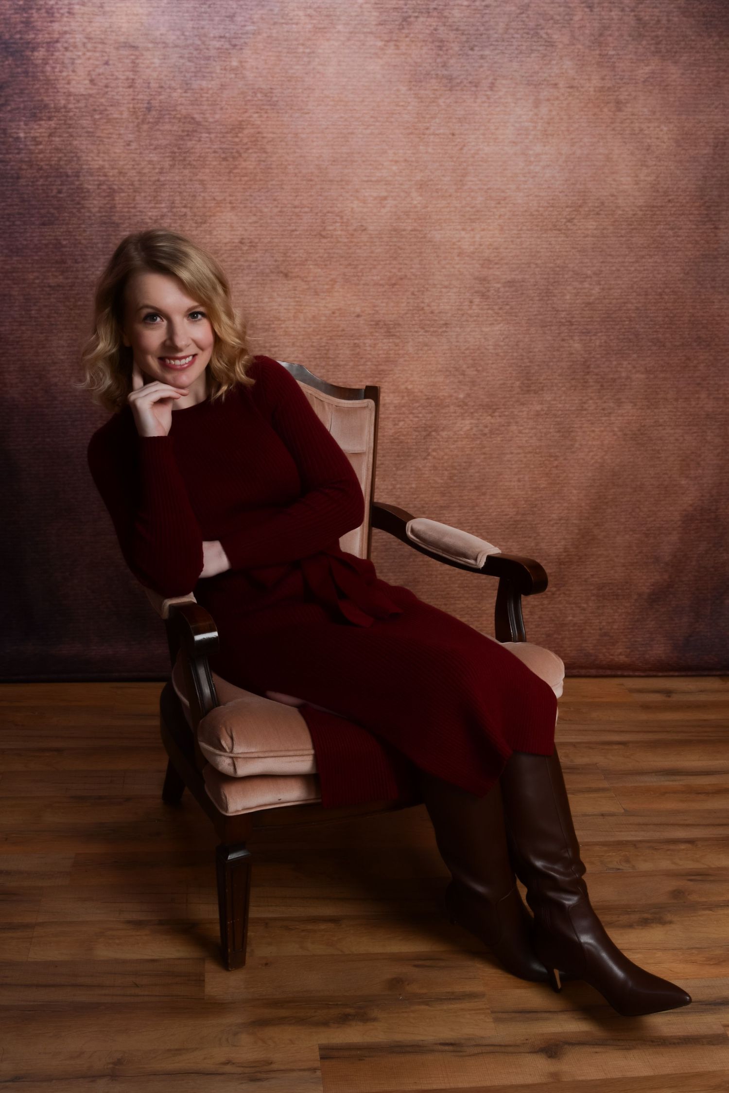 A person in a burgundy sweater dress and brown boots sits casually in a wooden chair against a textured brown backdrop.