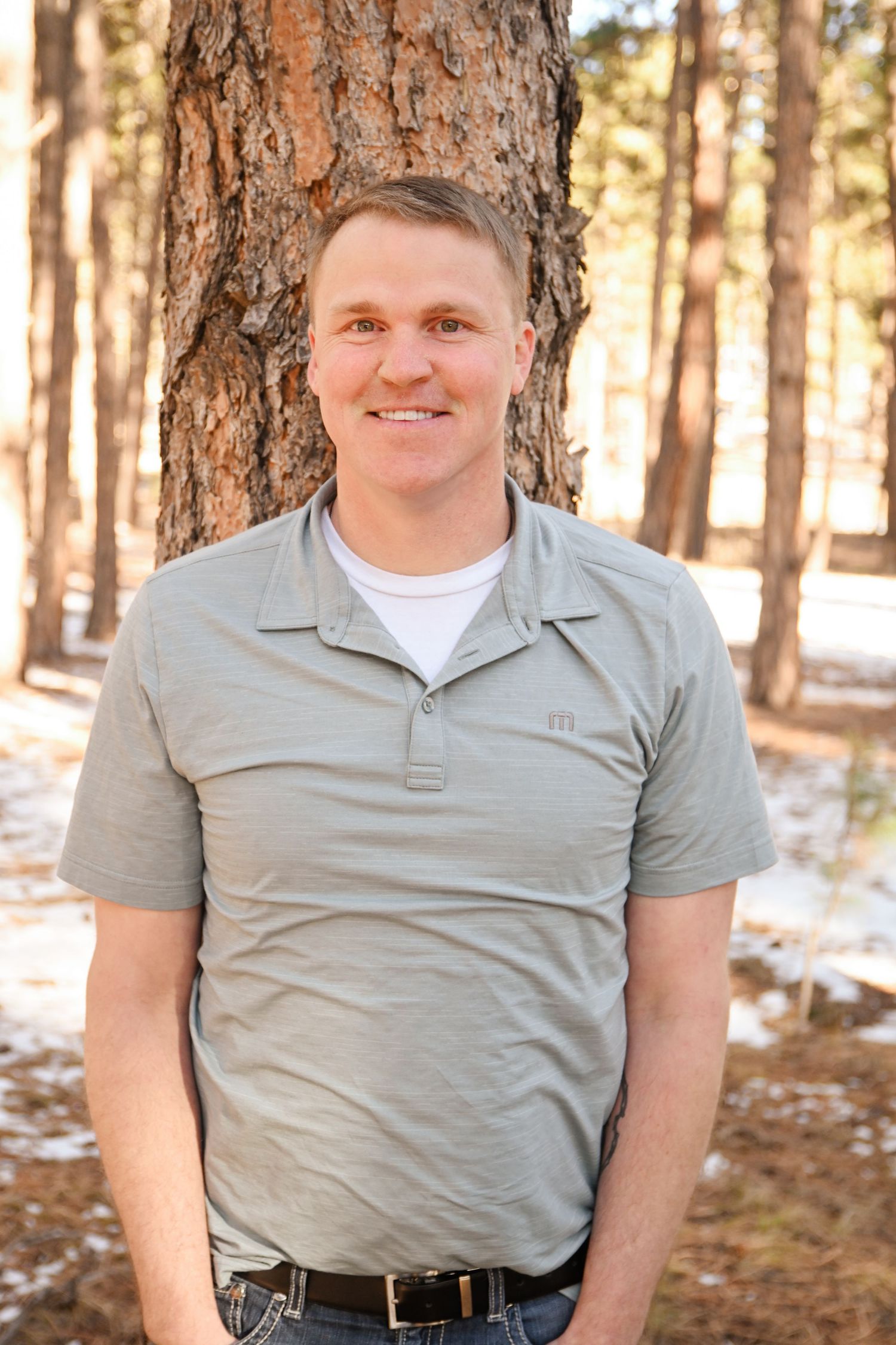 Person wearing a grey polo shirt poses against a pine tree in a snowy forest setting.