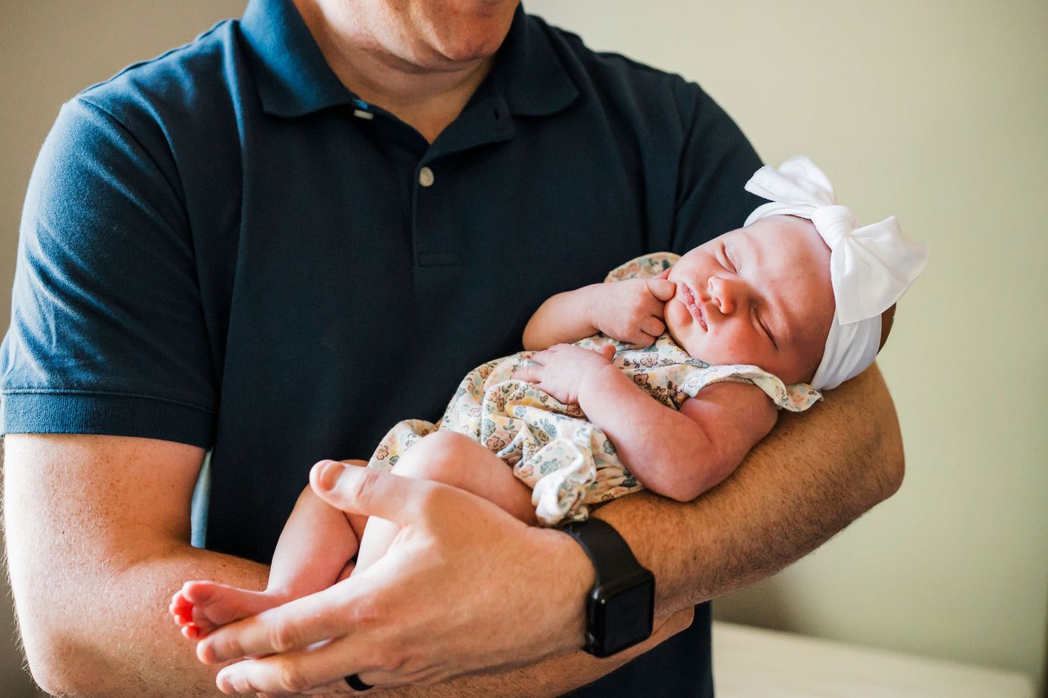 A newborn baby wearing a floral outfit and white bonnet sleeps peacefully in someone's arms.