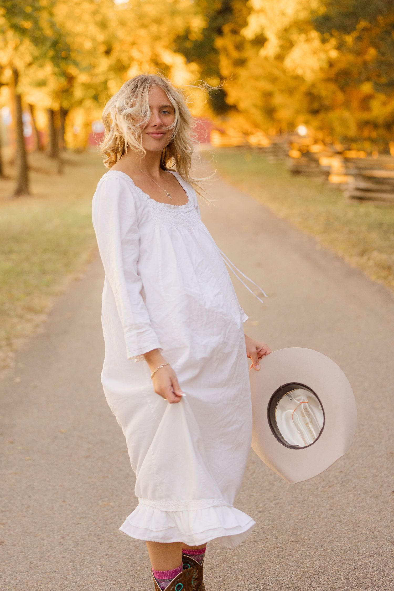 Person in a flowing white dress and boots stands on a path surrounded by golden autumn foliage in a park.