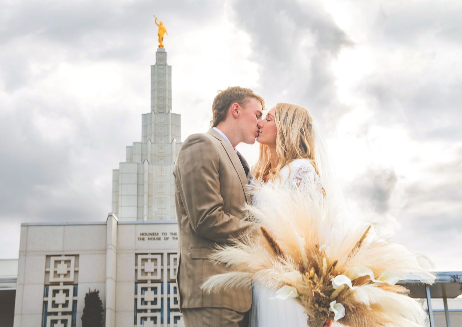 A romantic couple shares a kiss in front of the Idaho Falls Temple with a dramatic cloudy sky backdrop.