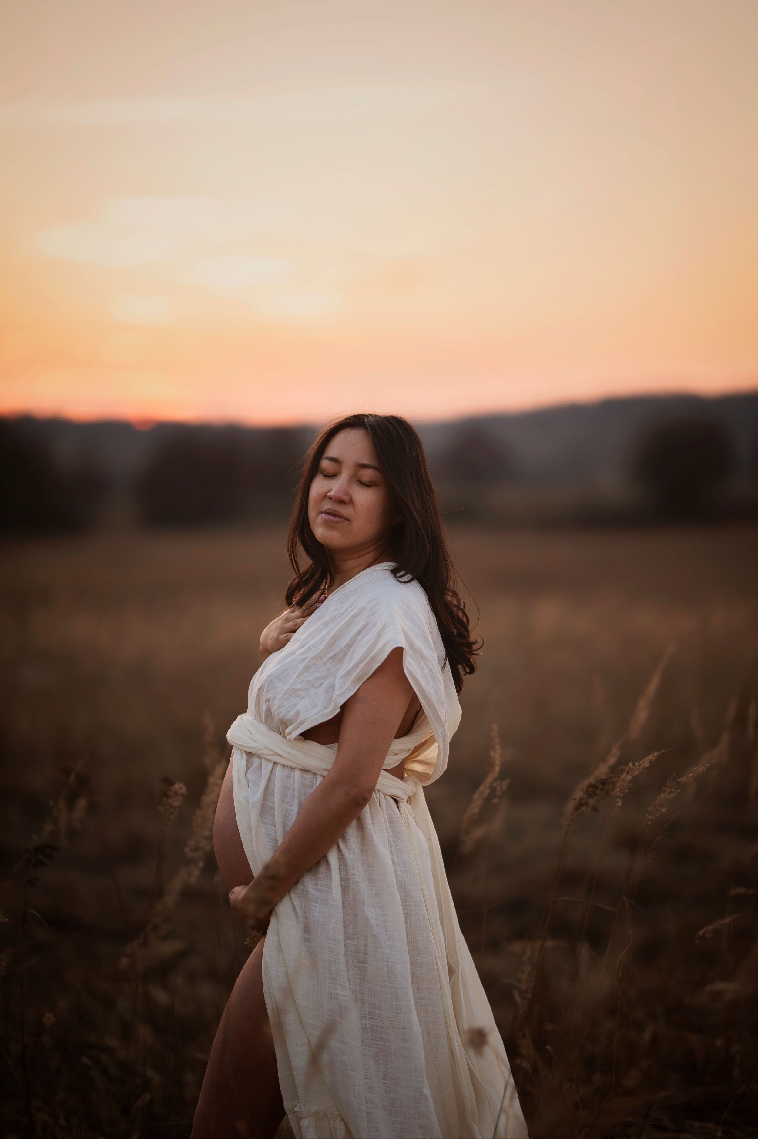 A pregnant woman in a flowing white dress stands in a field at sunset with golden light illuminating the scene.