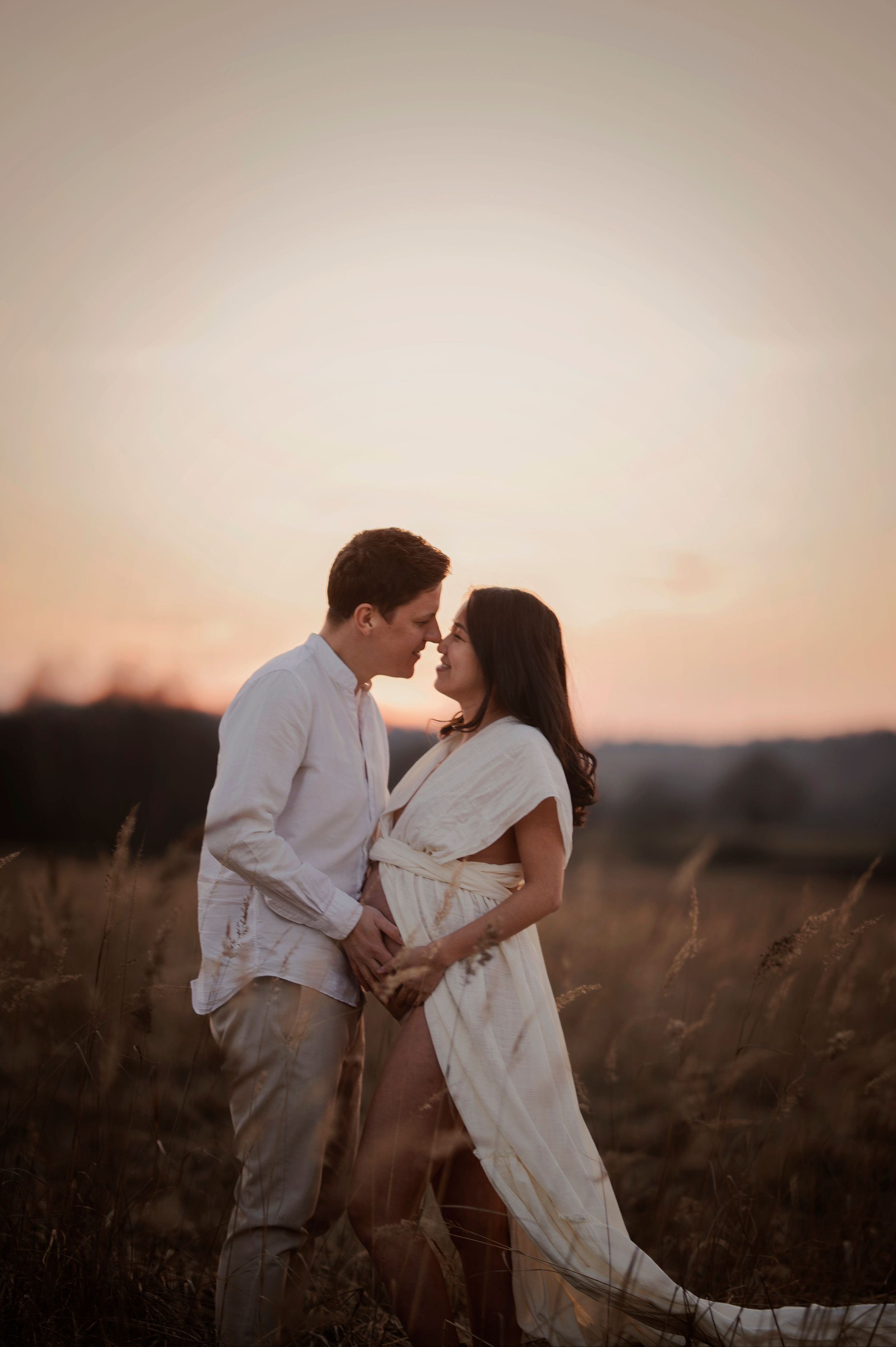 A romantic couple embraces in a field of tall grass during golden hour sunset with soft warm lighting.