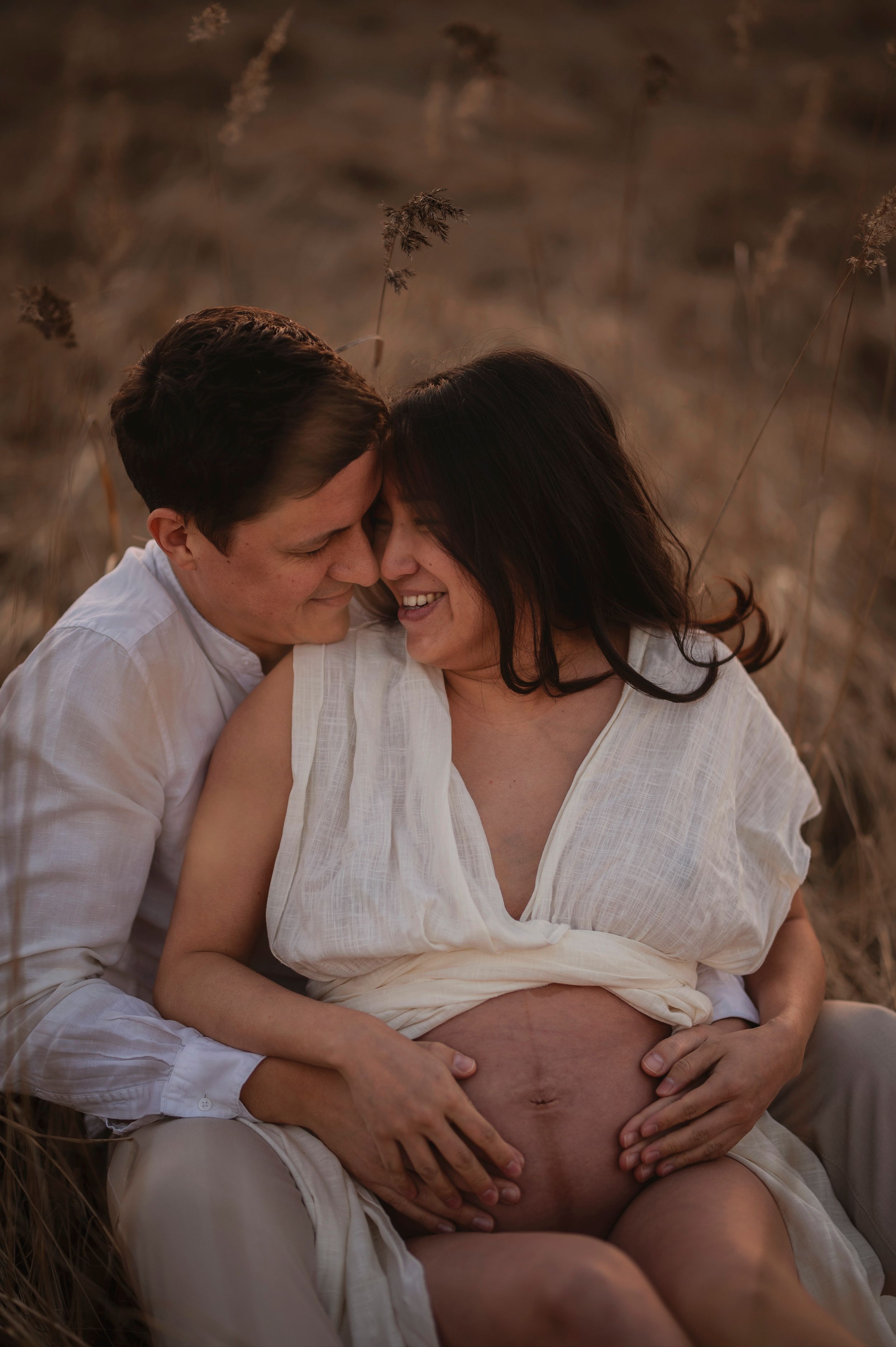 A tender maternity photo shows a couple in white clothing embracing in a sun-lit field while touching the pregnant belly.
