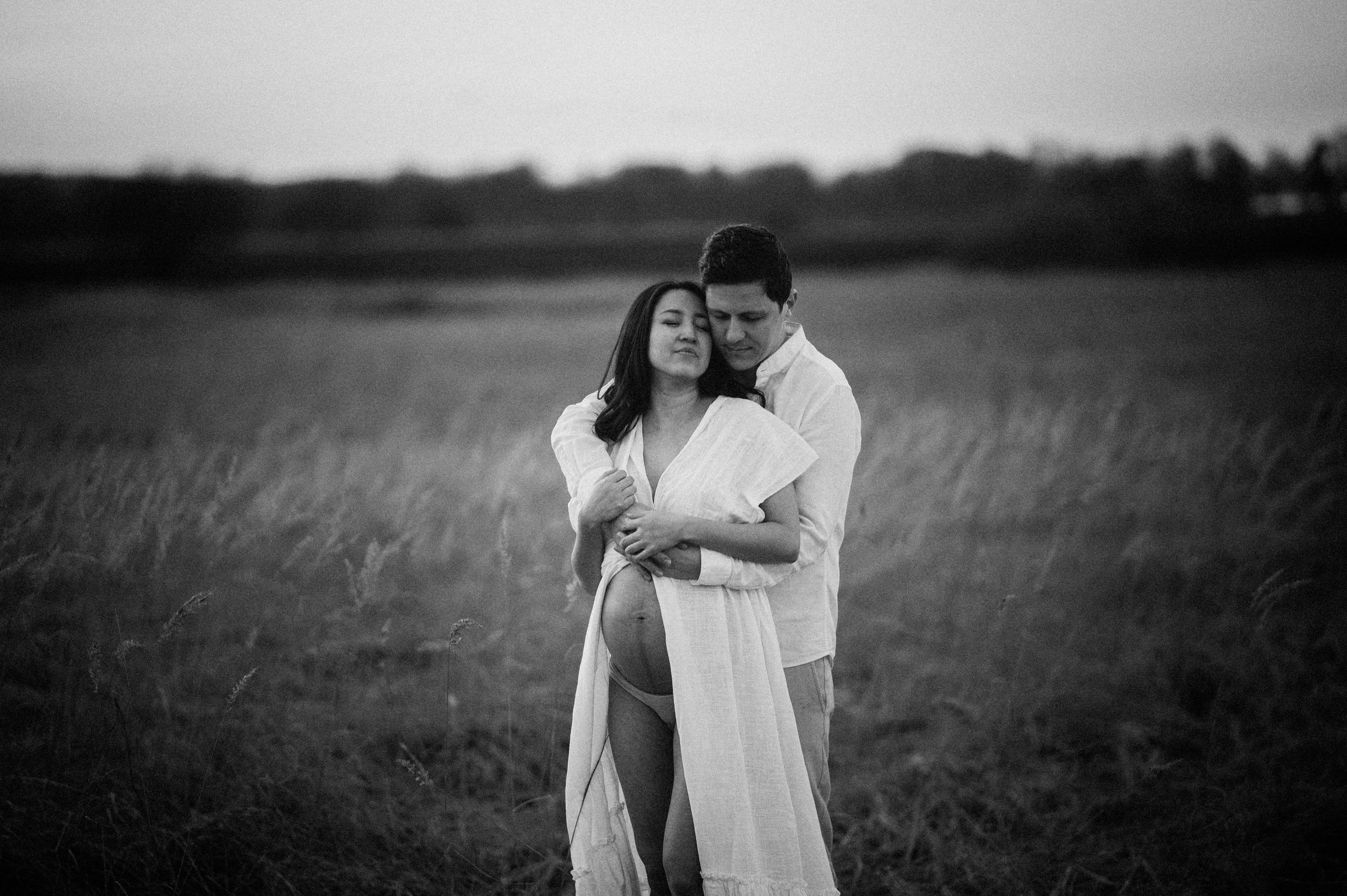 A romantic black and white photo of a couple embracing in a serene field of tall grass at dusk.