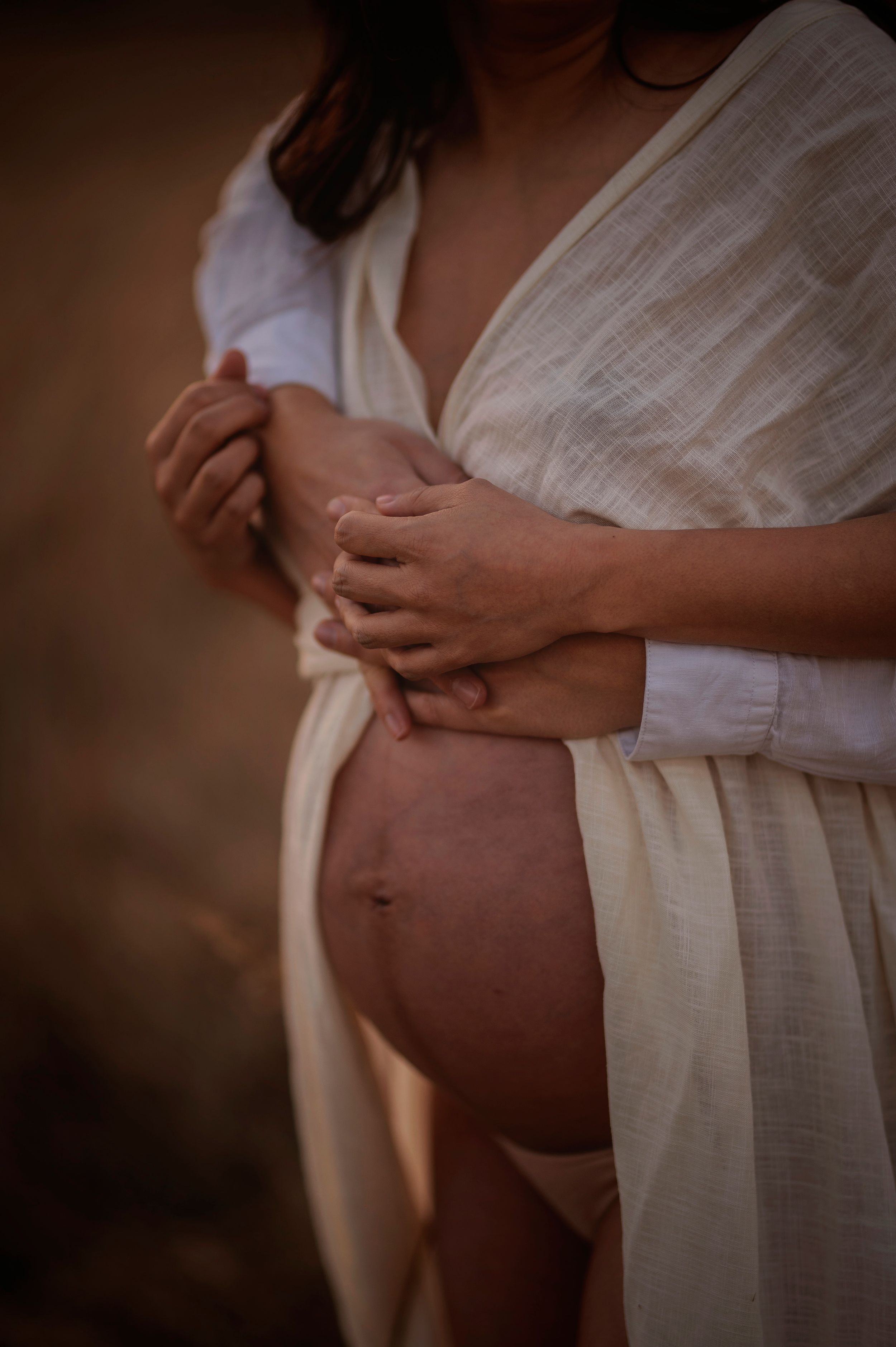 A pregnant woman in a white flowing dress holds her belly in warm natural lighting during a maternity photoshoot.
