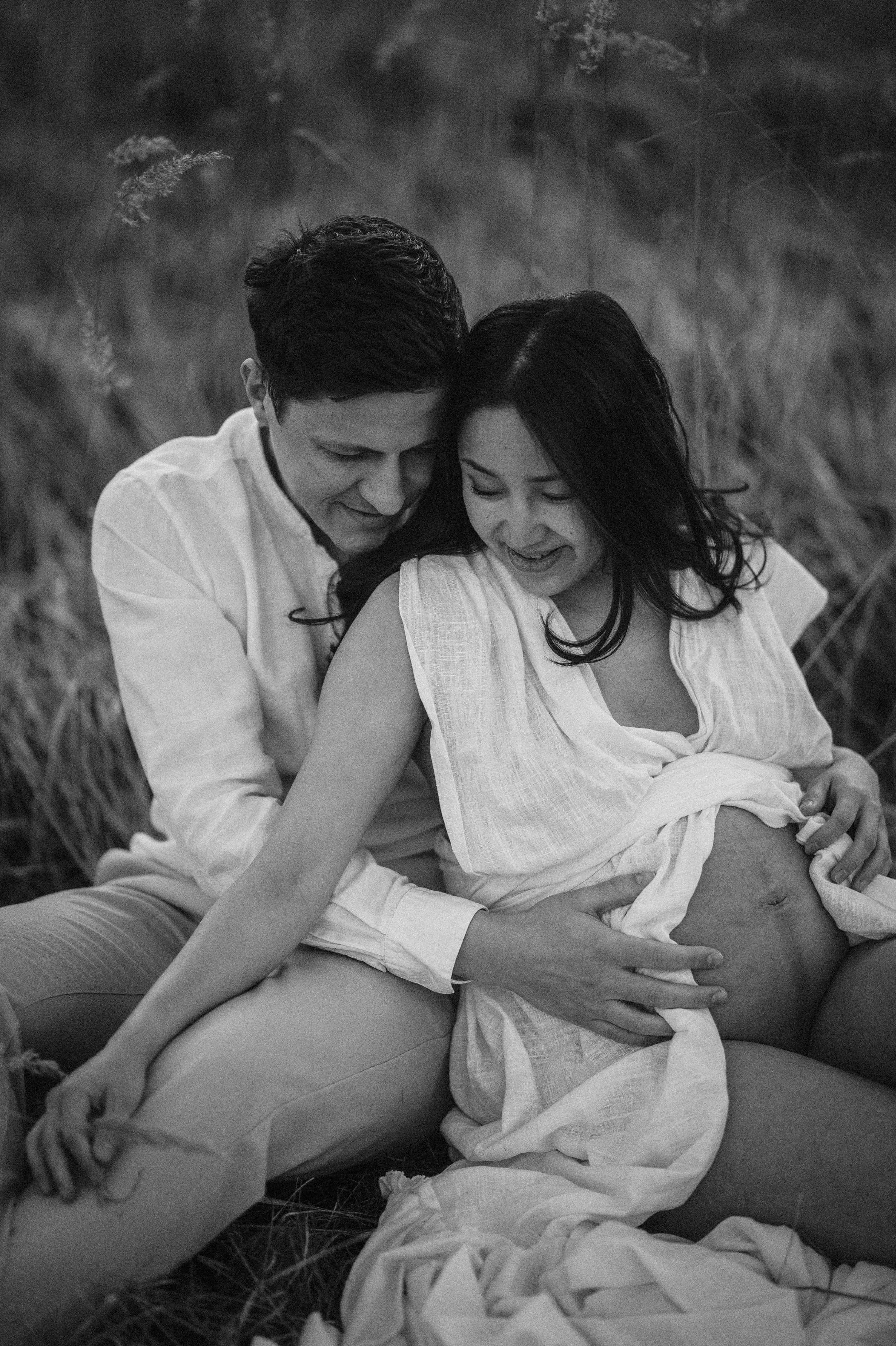 An intimate black and white portrait of a couple in white clothing embracing on a blanket in a grassy field.