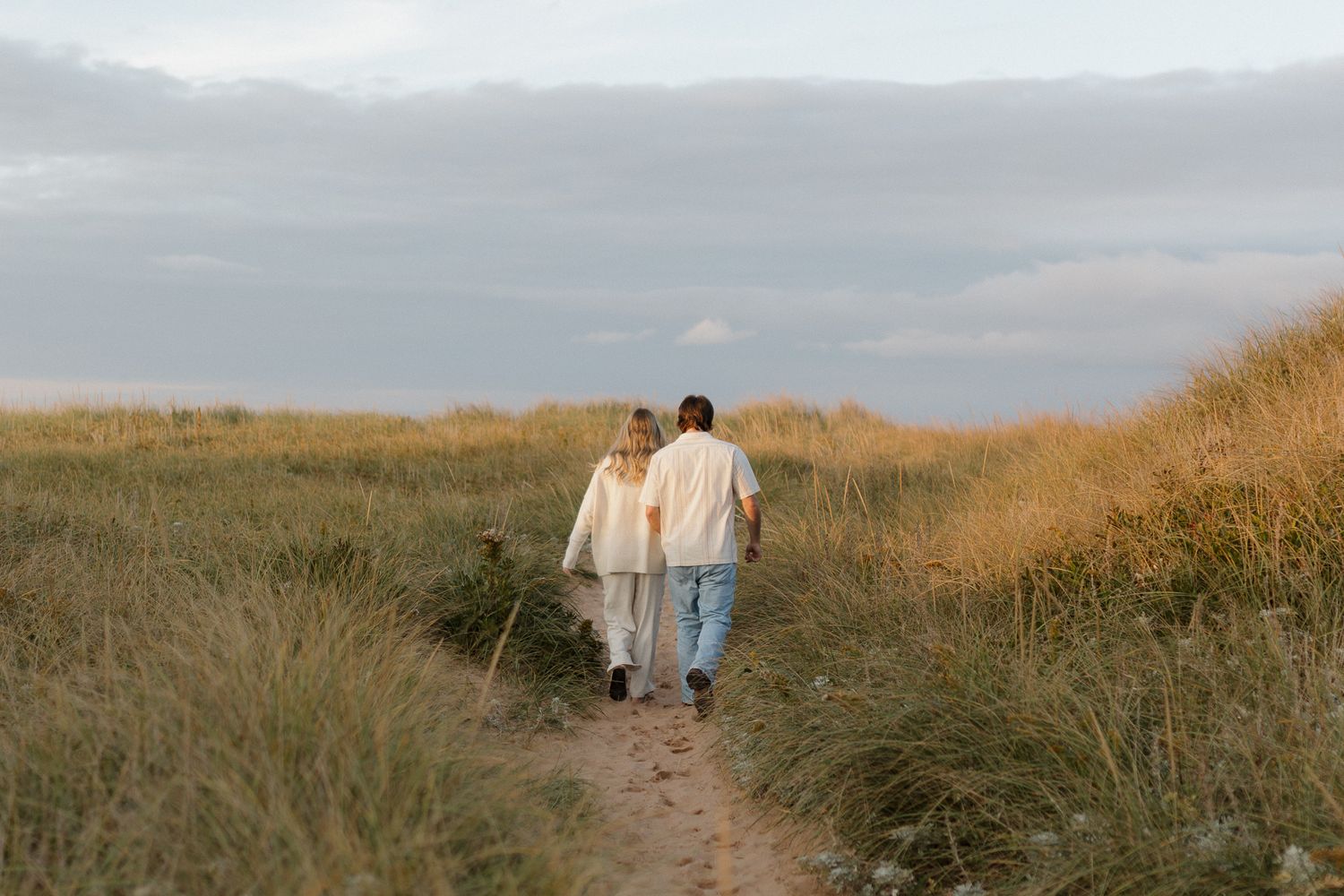 Couple walking together on dirt path through tall grass at sunset.